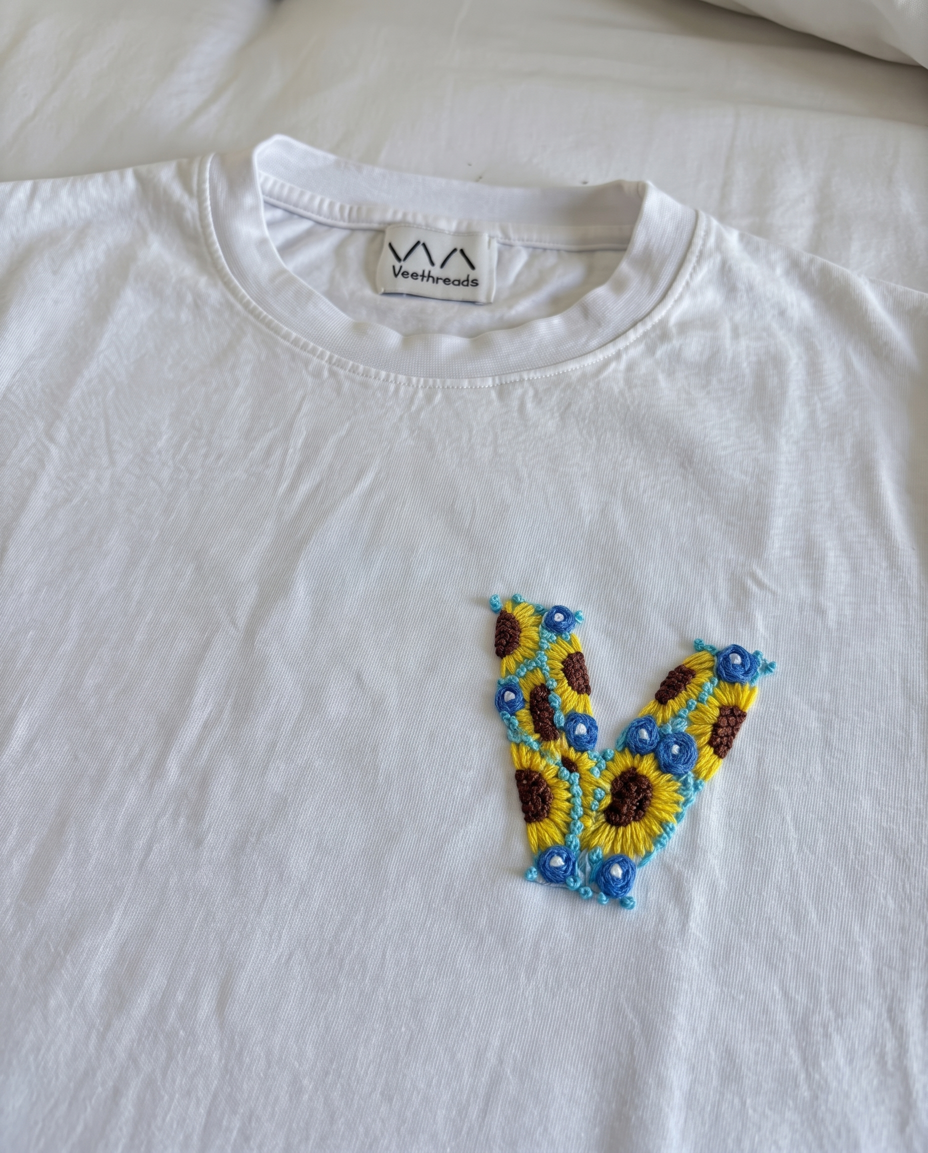 White cotton tee with an initial hand-embroidered as sunflowers and small blue flowers, by Vee Threads
