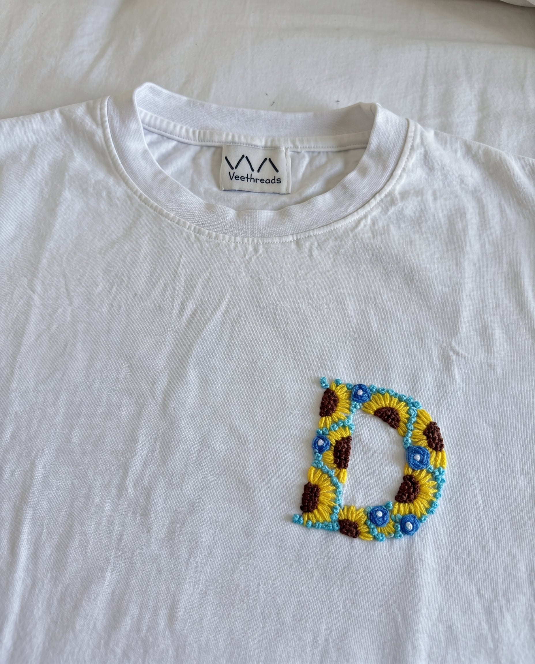 Sunflower and blue-flower hand-embroidered letter on a white cotton tee, by Vee Threads