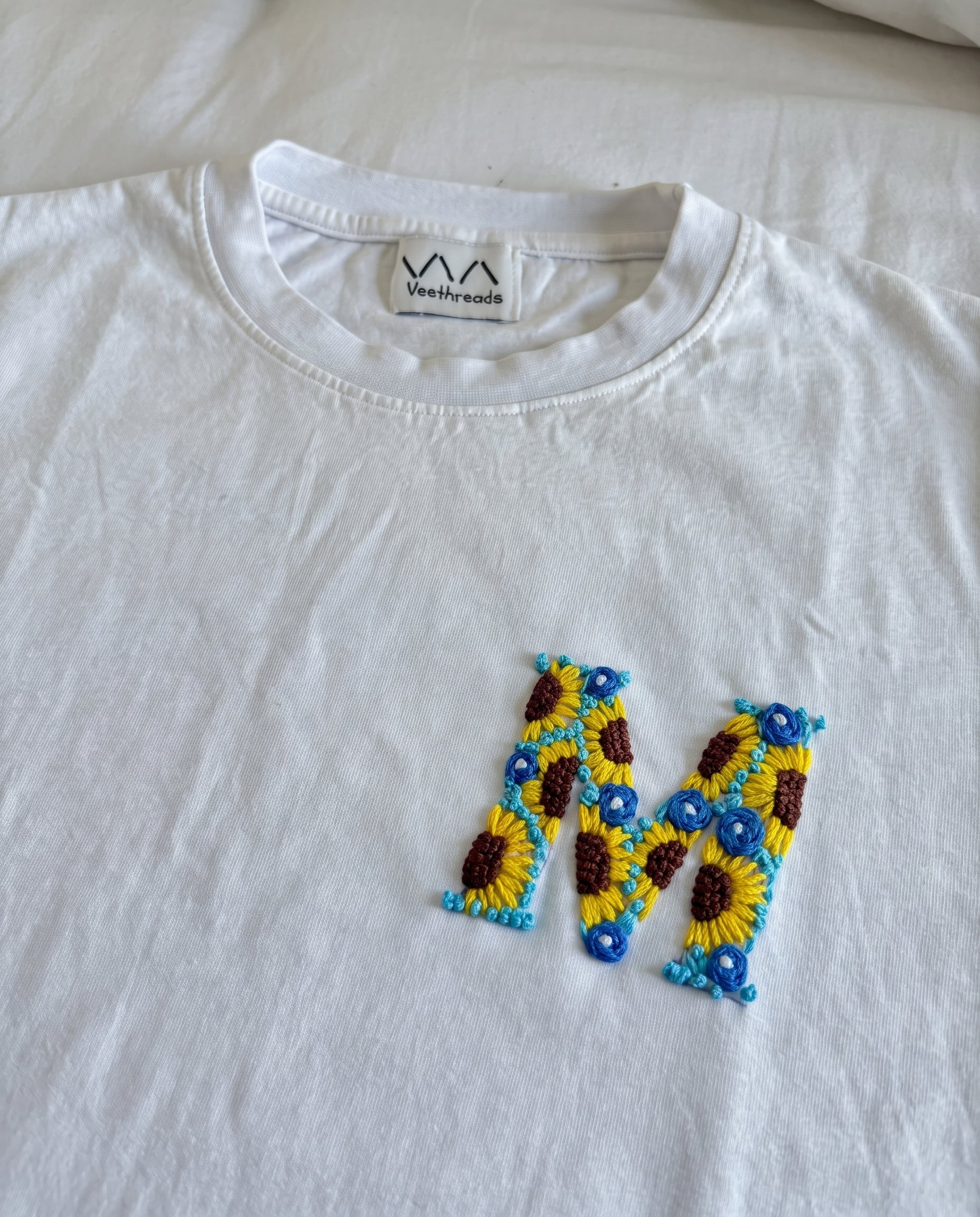 White cotton tee with an initial hand-embroidered as sunflowers and small blue flowers, by Vee Threads