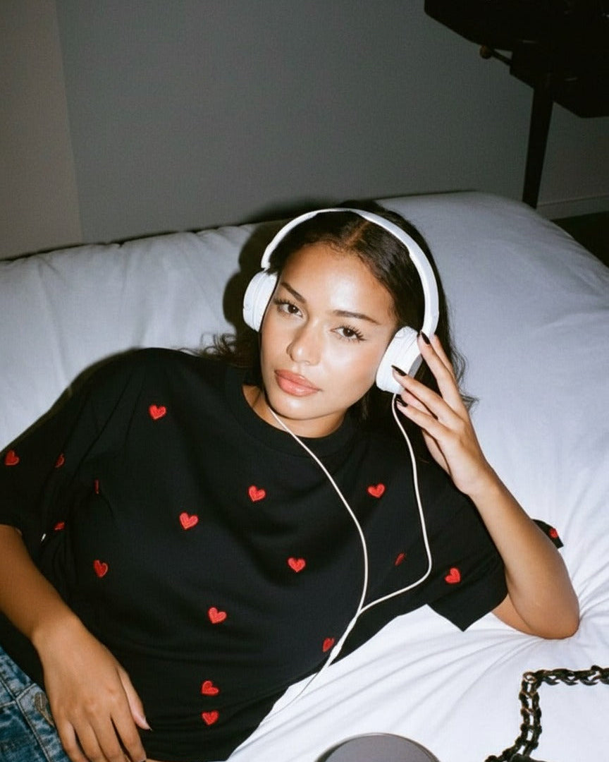 Woman wearing white red heart hand embridered on it headphones and a black shirt with red heart patterns, sitting on a bed.