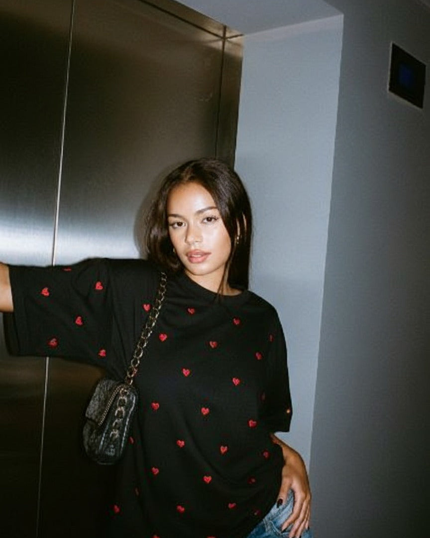 Woman wearing a black shirt with red heart patterns embroidered and holding a handbag indoors.