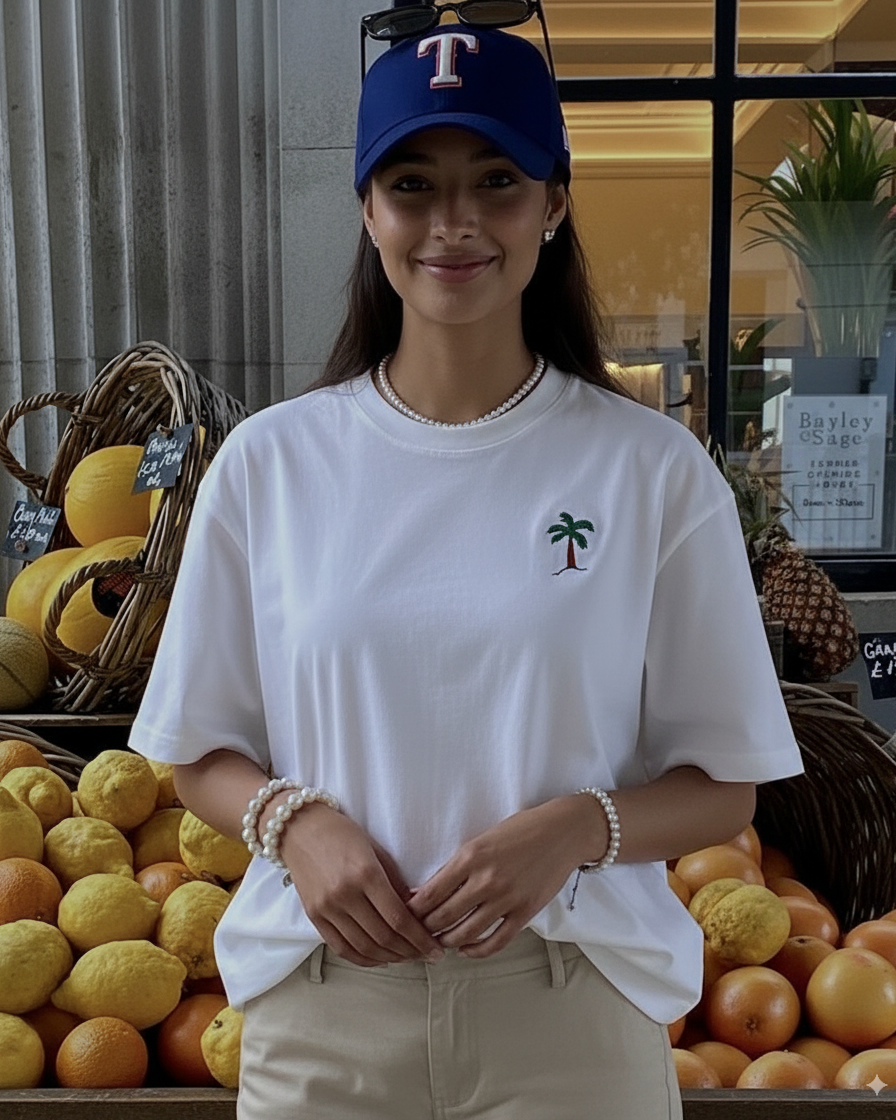 Person wearing a white t-shirt with a palm tree design hand embroidered , blue cap, and beige pants standing in front of a fruit stand.