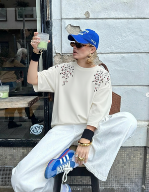 Person wearing a blue cap and white sweatshirt with floral patterns, sitting outside a building.