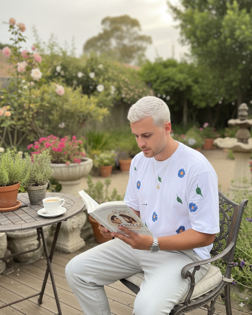 Man wearing embroidered floral tshirt reading a book in a garden setting with plants and a fountain.