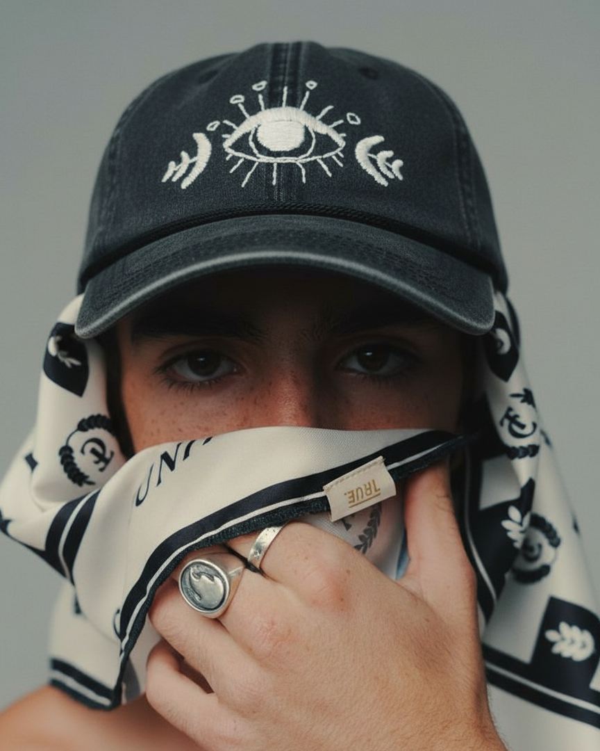 Person wearing a black cap with white designs embroidered and a patterned scarf, holding a phone.