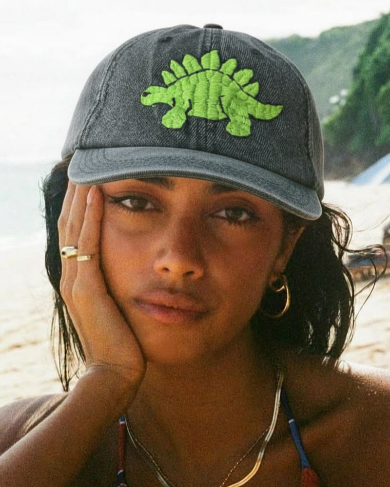 Person wearing a cap with a green dinosaur design embroidered on a beach