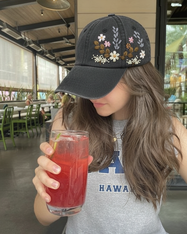 Person wearing a floral embroidered cap and holding a glass of red drink in an outdoor setting.