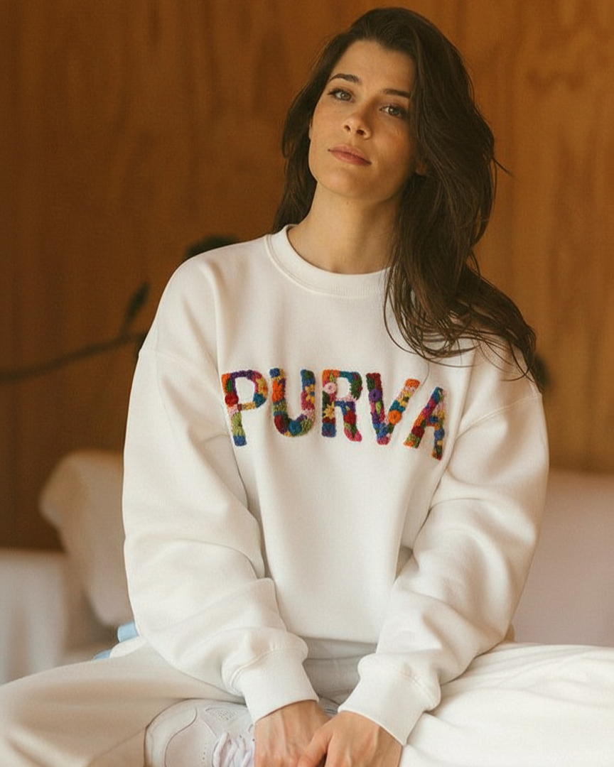 Woman wearing a white sweatshirt with 'PURVA' embroidered in colorful letters, sitting against a wooden wall.