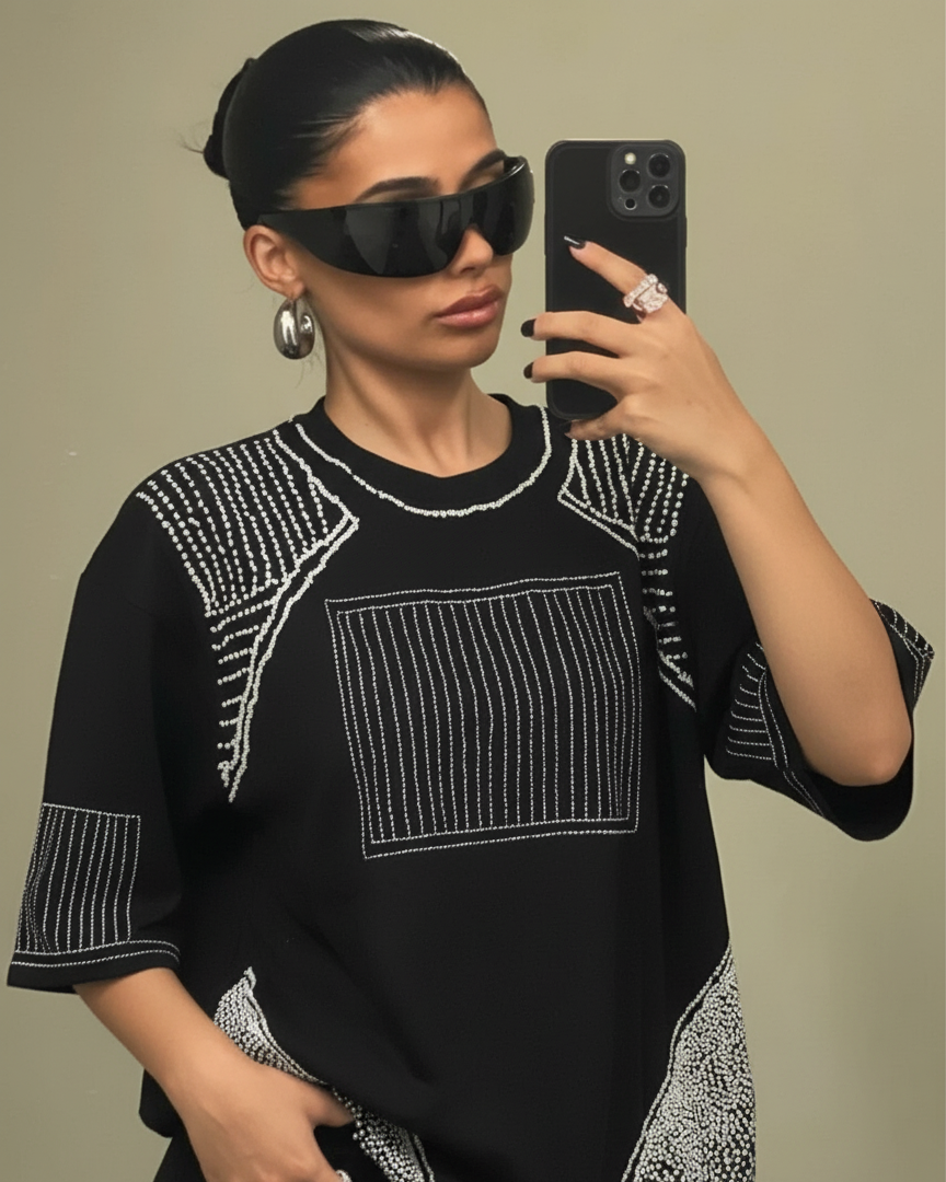 Woman wearing a black top with white patterns embroidered , taking a mirror selfie.