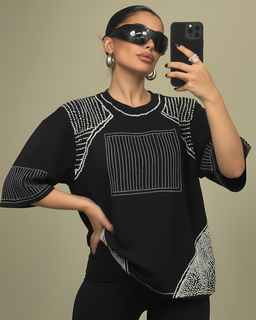 Woman wearing a embroidered tshirt taking a mirror selfie wearing a black outfit with white patterns on a beige background