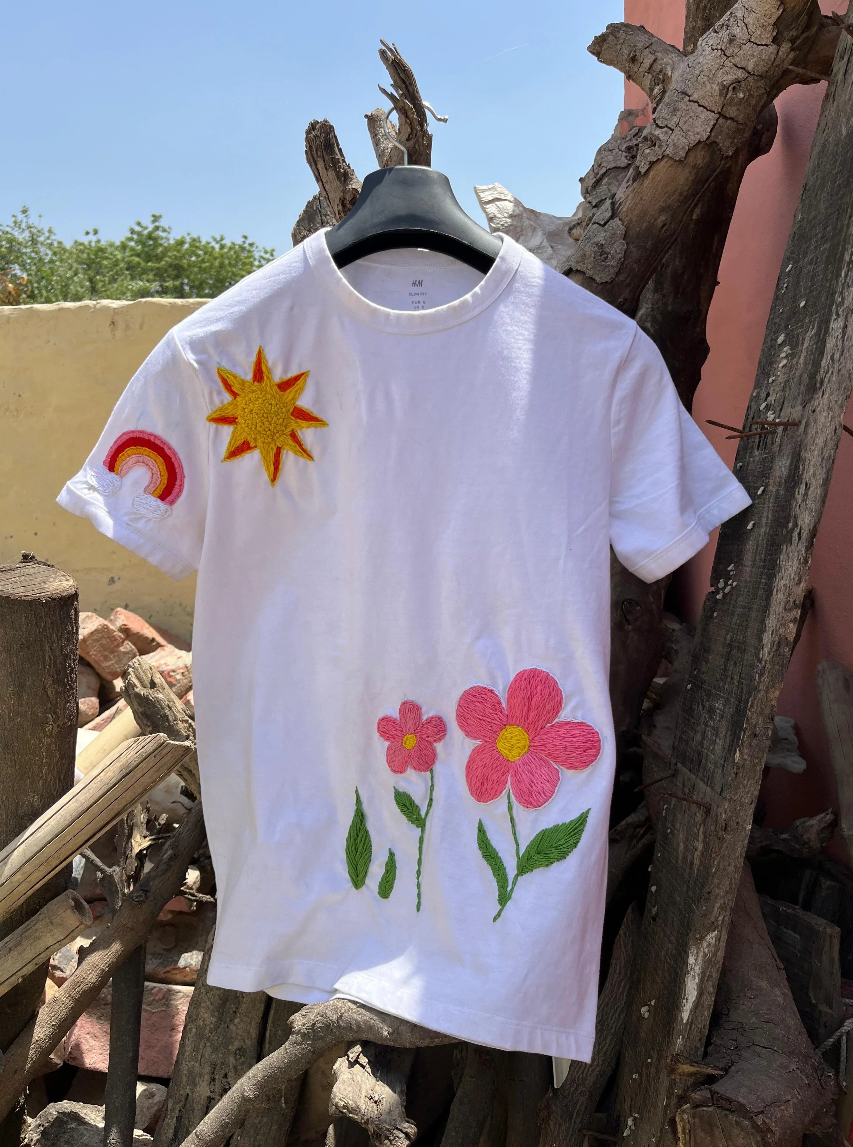 White t-shirt with colorful floral embrloidery designs hanging on a wooden structure.