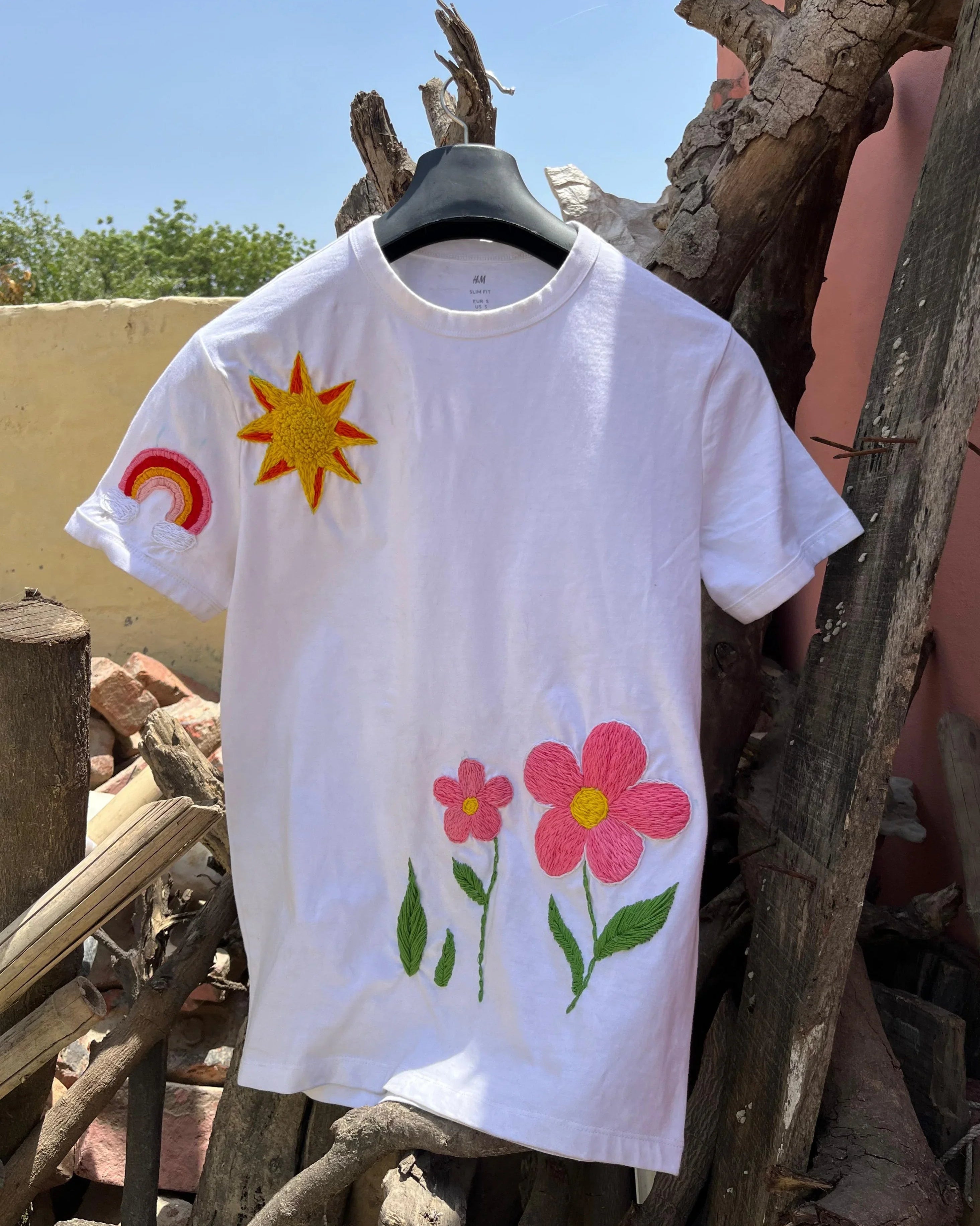 White t-shirt with colorful floral embrloidery designs hanging on a wooden structure.