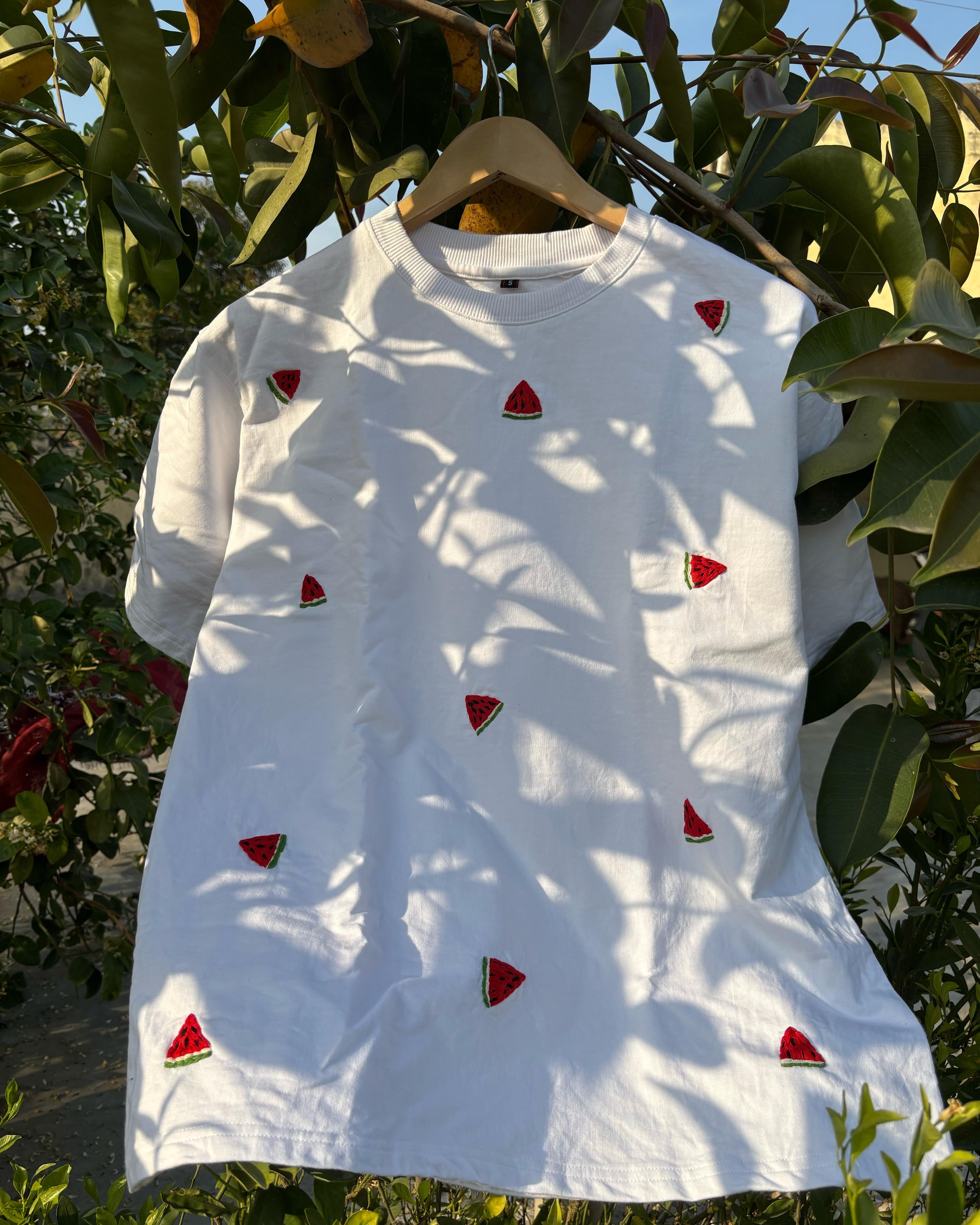 A white oversized t-shirt with a hand-embroidered watermelon motif, displayed on a hanger outdoors.