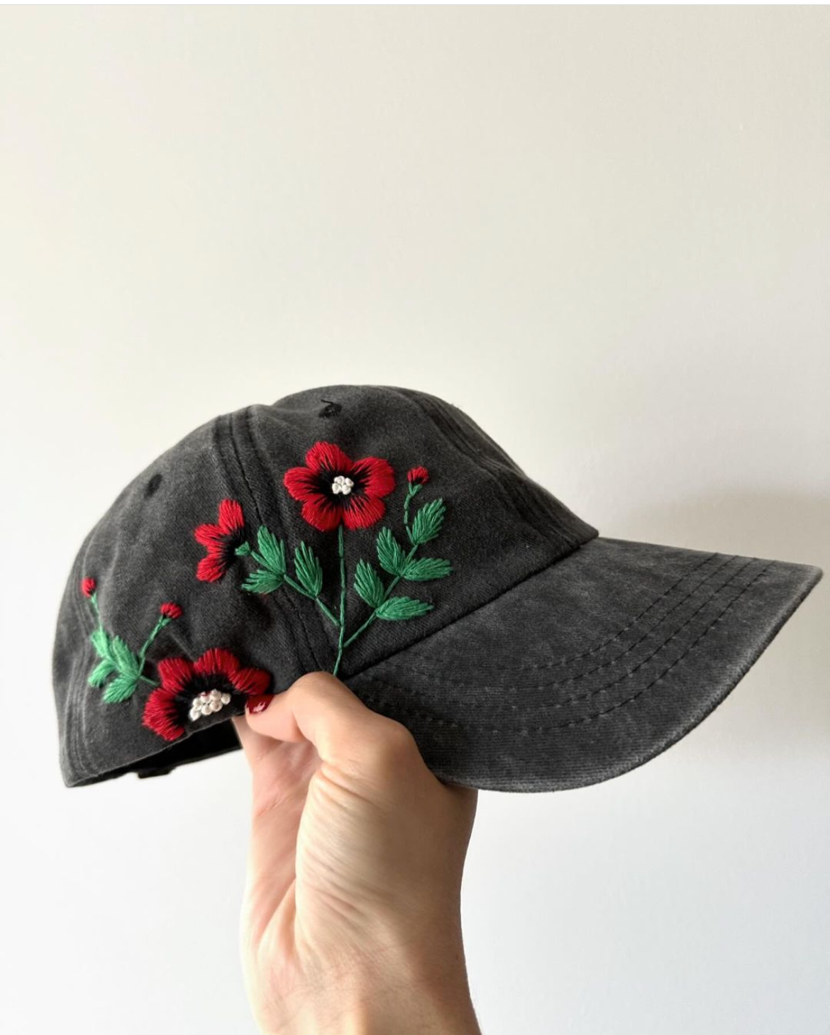 A hand holding a gray denim cap with red floral embroidery on the front.