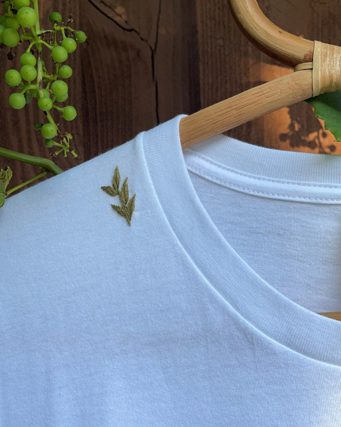 Embroidered tee leaf motif — light blue cotton tee with hand-embroidered green leaf design on wooden hanger against wooden background