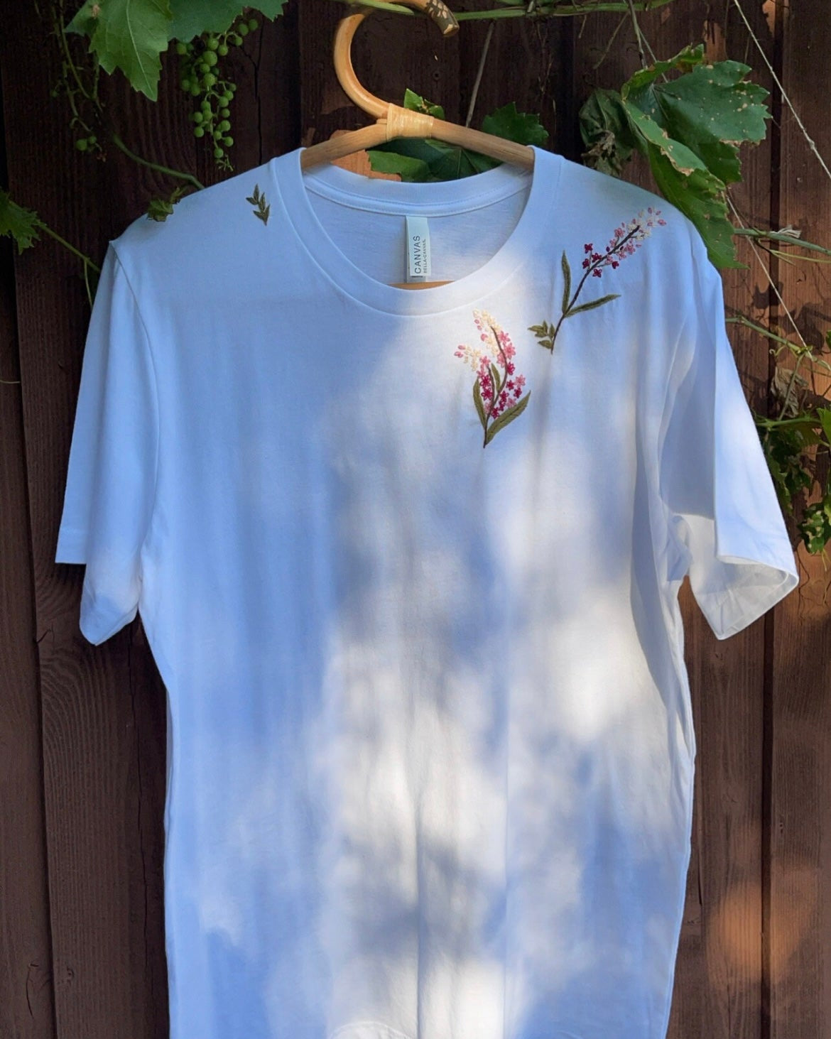 Embroidered tee floral garden — white cotton tee with hand-stitched floral embroidery on wooden fence with greenery background