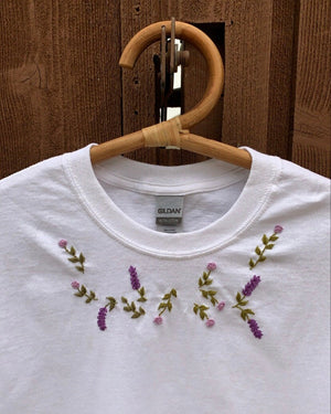 White oversized t-shirt with hand-embroidered floral patterns on the front, displayed on a wooden hanger.