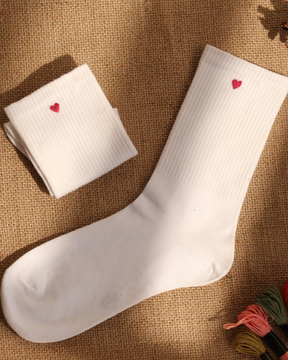 A pair of white hand embroidered socks with red heart designs, displayed on a burlap surface with pine cones and yarn nearby.