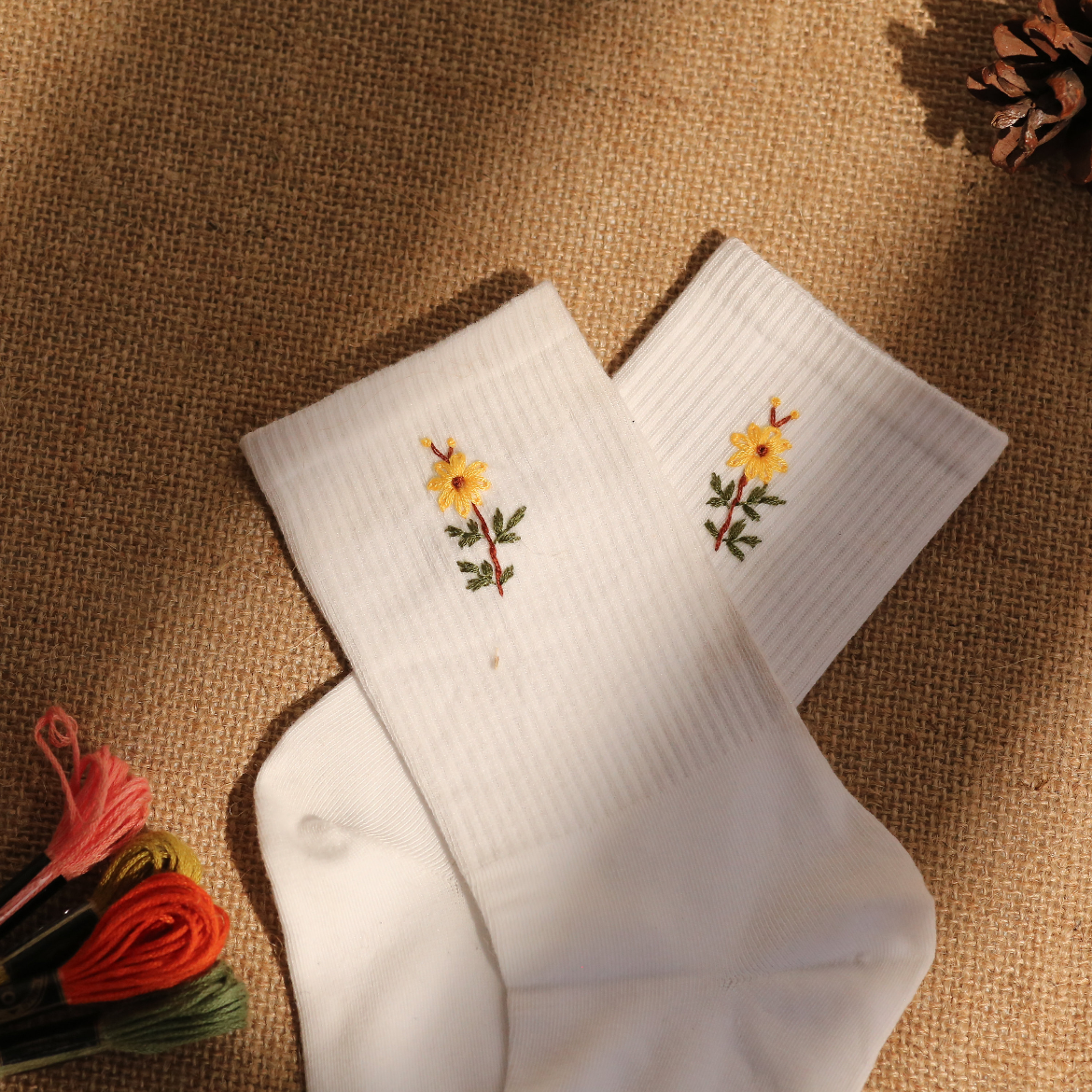 A pair of white hand-embroidered socks with sunflower designs, laid out on a burlap surface with a few embroidery threads and pine cones nearby.