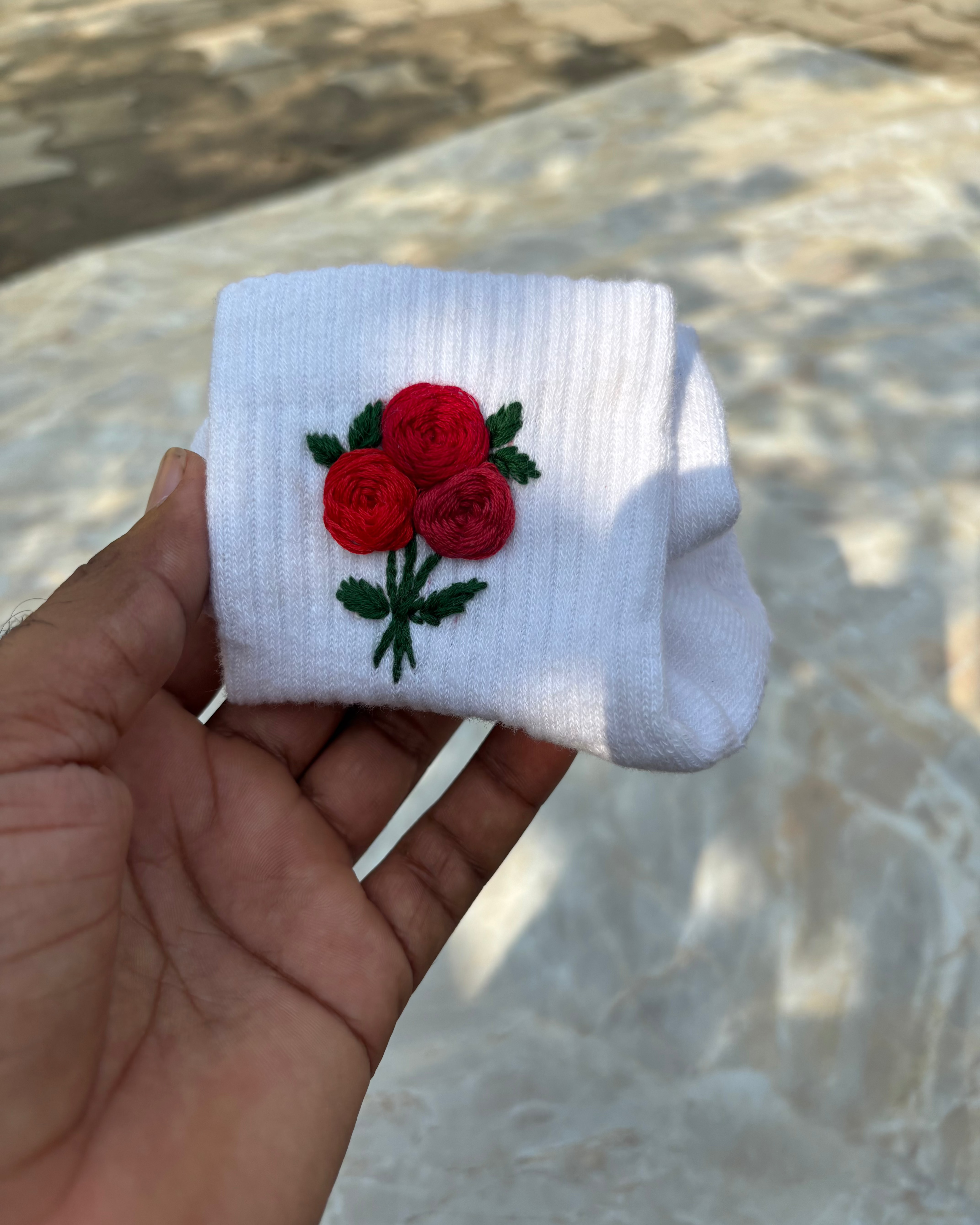 A pair of white hand-embroidered socks with a rose design held in a person's hand, with a blurred background.