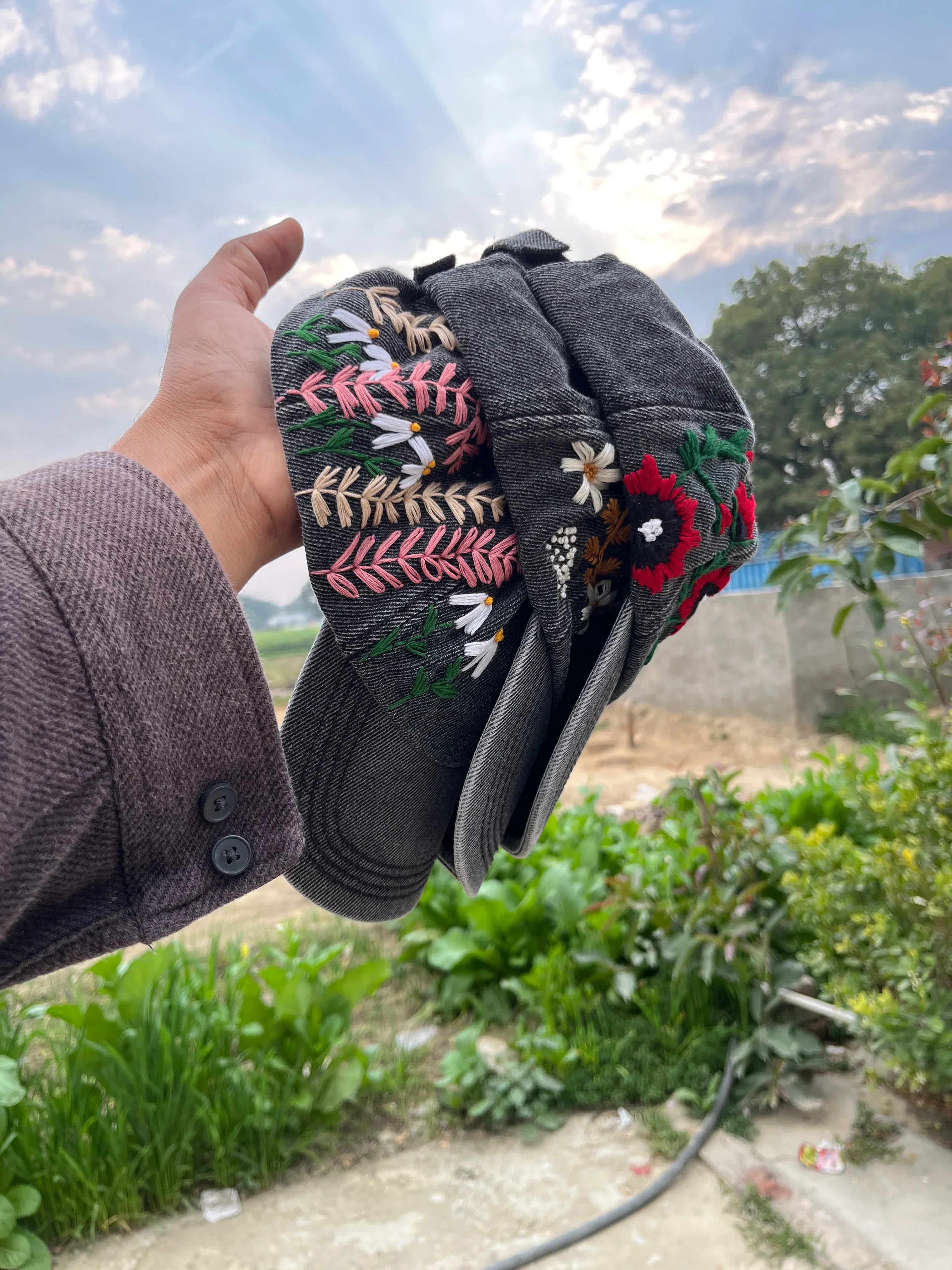 Hand holding a black cap with colorful floral embroidery against a blurred outdoor background