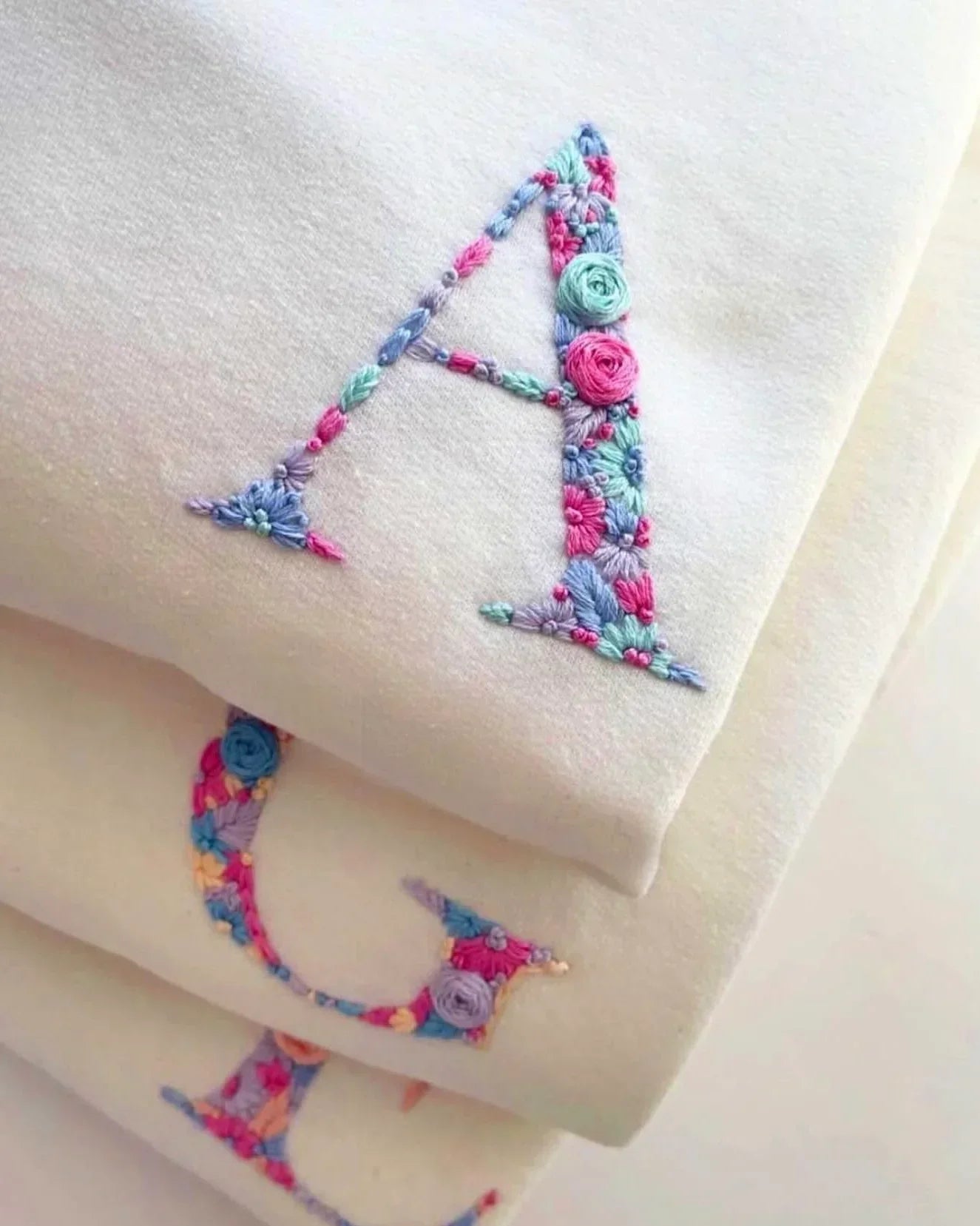 Flatlay of folded white custom initials t-shirt with black embroidery on chest, placed beside sunglasses and a phone.