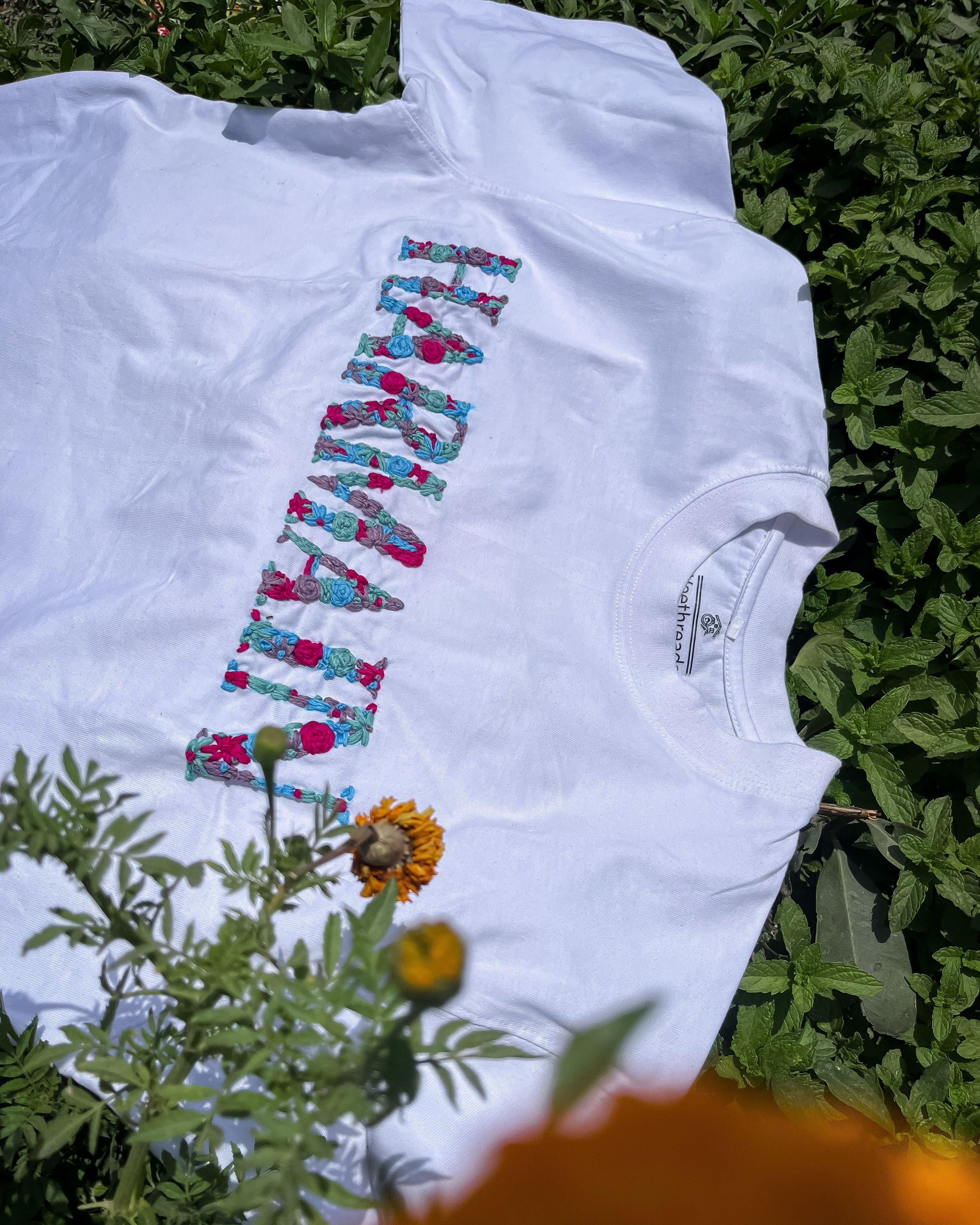 Custom white tee featuring embroidered name across chest, displayed with gifting accessories like ribbon and note card.