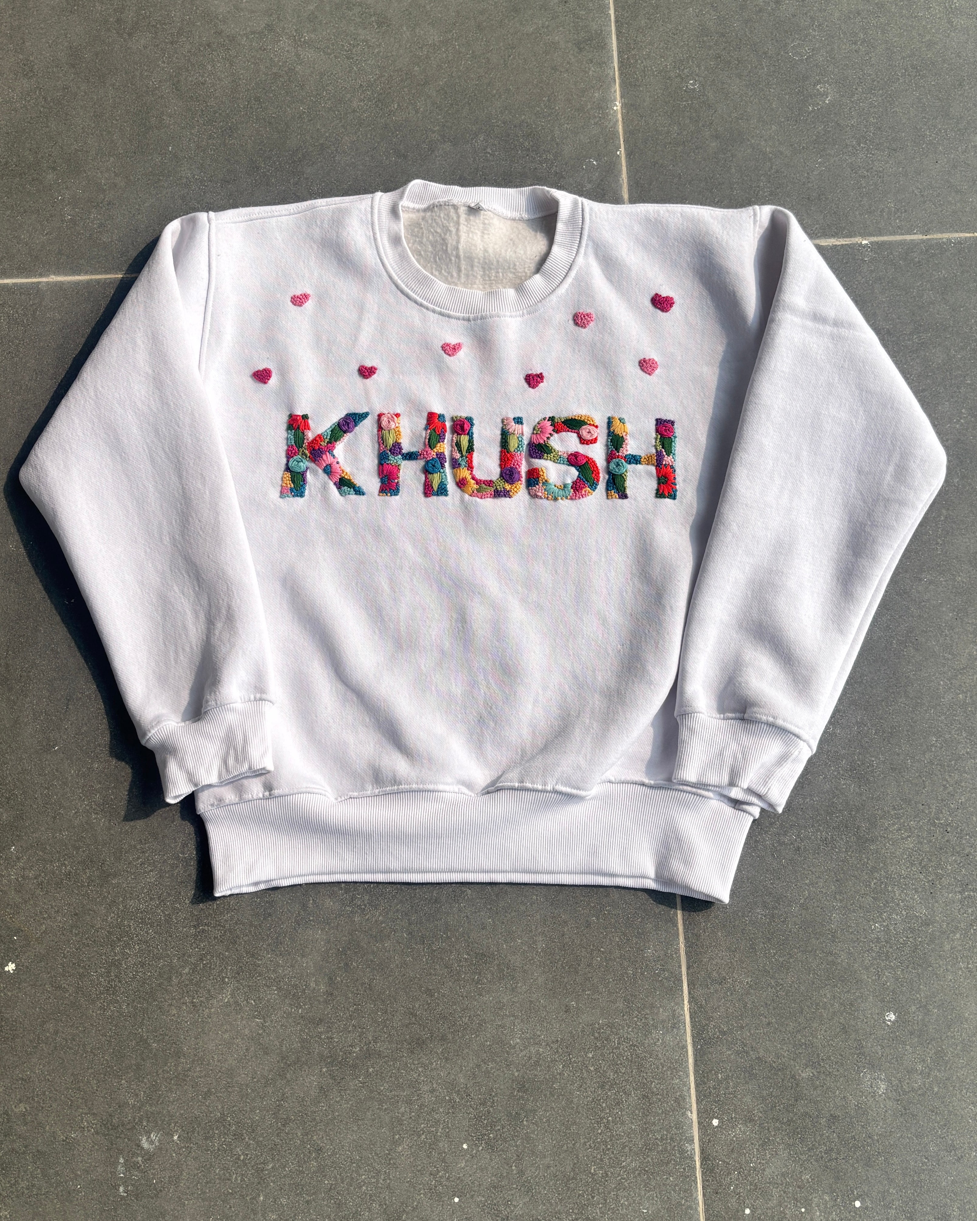 White sweatshirt with custom name hand embroidered and heart patterns on a gray floor.