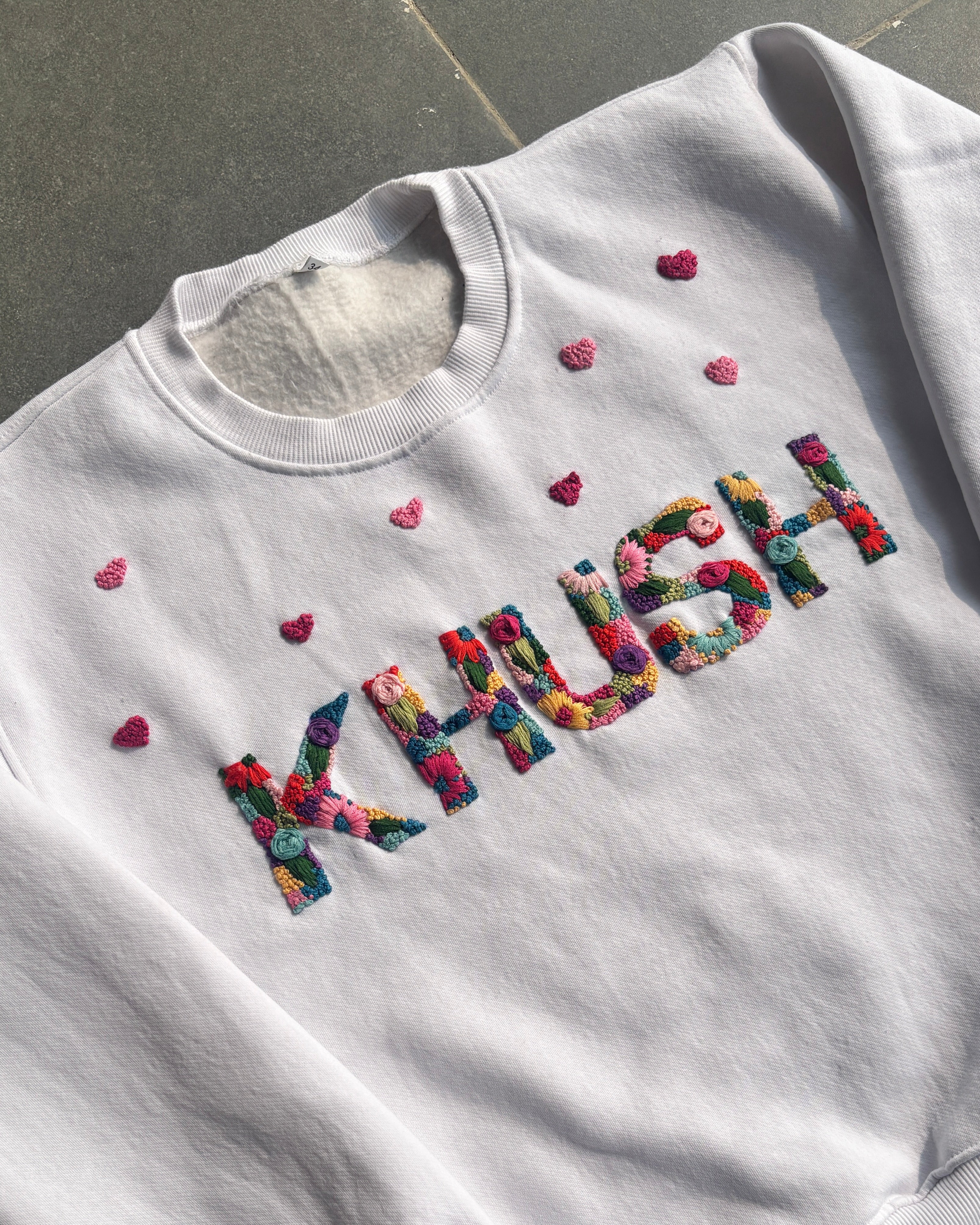 White sweatshirt with colorful 'Kawaii' custom name hand embroidered and small hearts on a dark surface