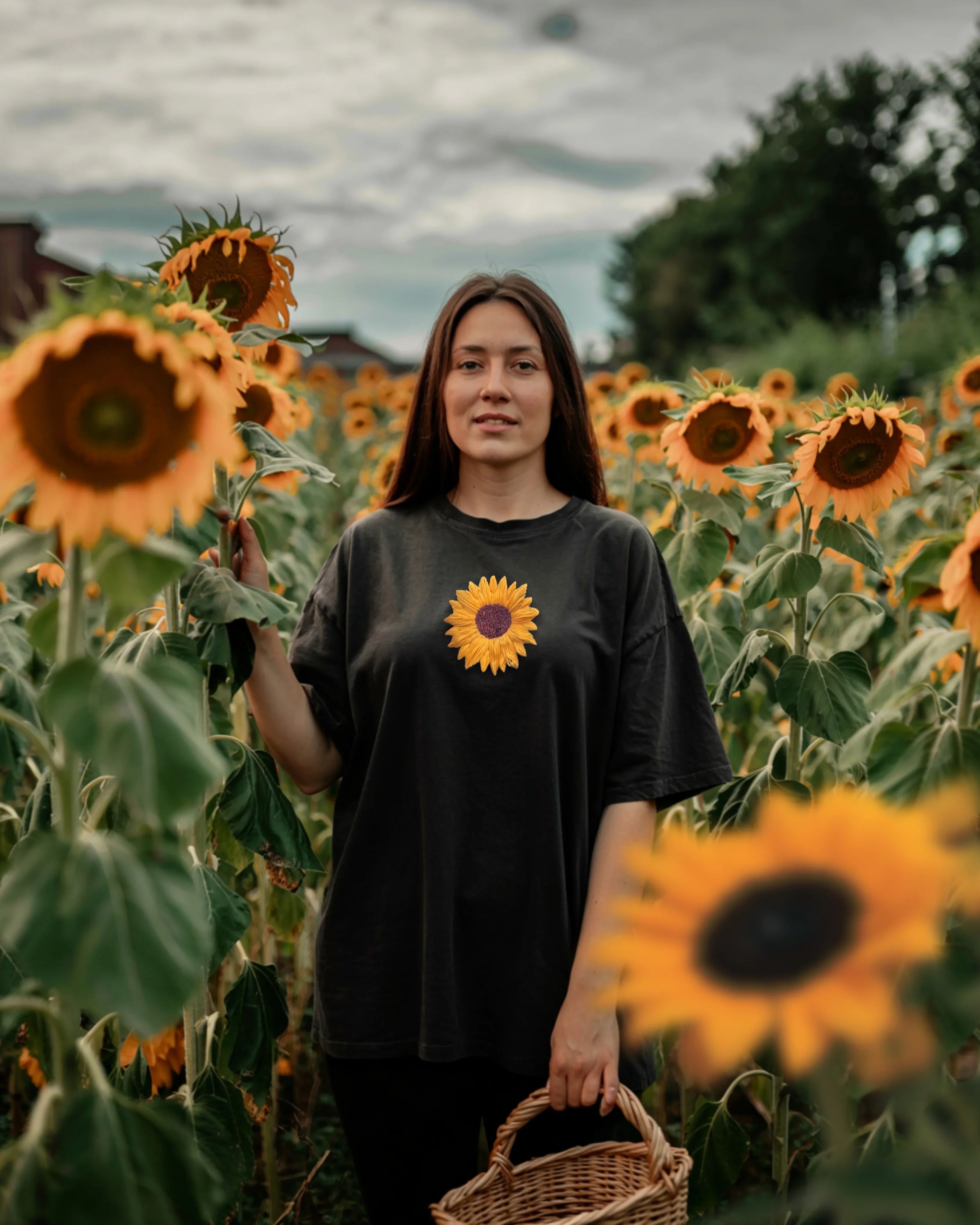 Woman standing in a sunflower field wearing a black t-shirt with a sunflower design embroidered on it.