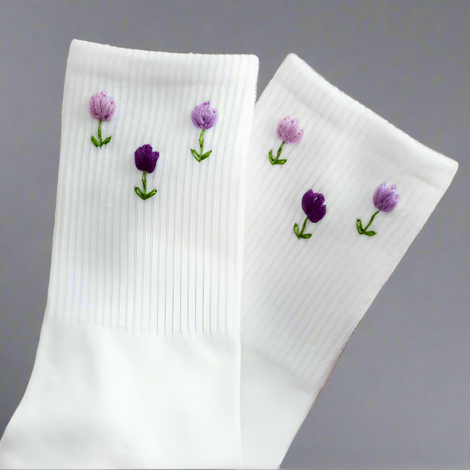 A pair of white embroidery socks with purple tulip flowers designs on the top side, displayed against a grey background.