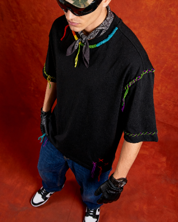 Person wearing a black shirt with colorful patterns, blue jeans, and black gloves on a red background