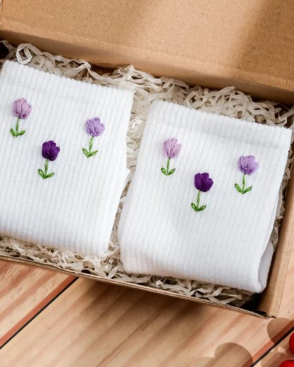 Two white embroidered socks with purple flowers in a cardboard box on a wooden surface.