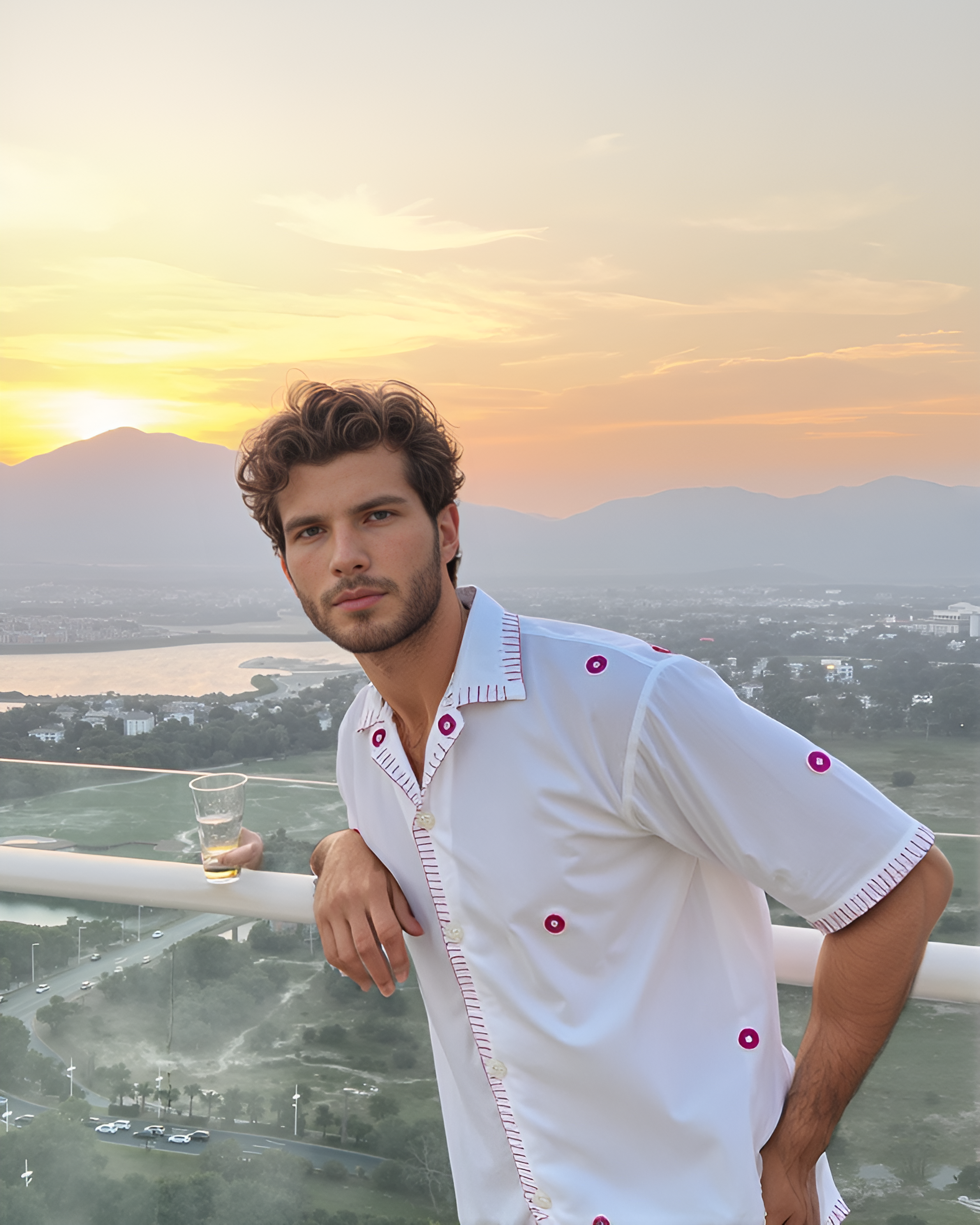 Man in a white shirt with mirror work embroidery standing on a balcony overlooking a scenic landscape at sunset.