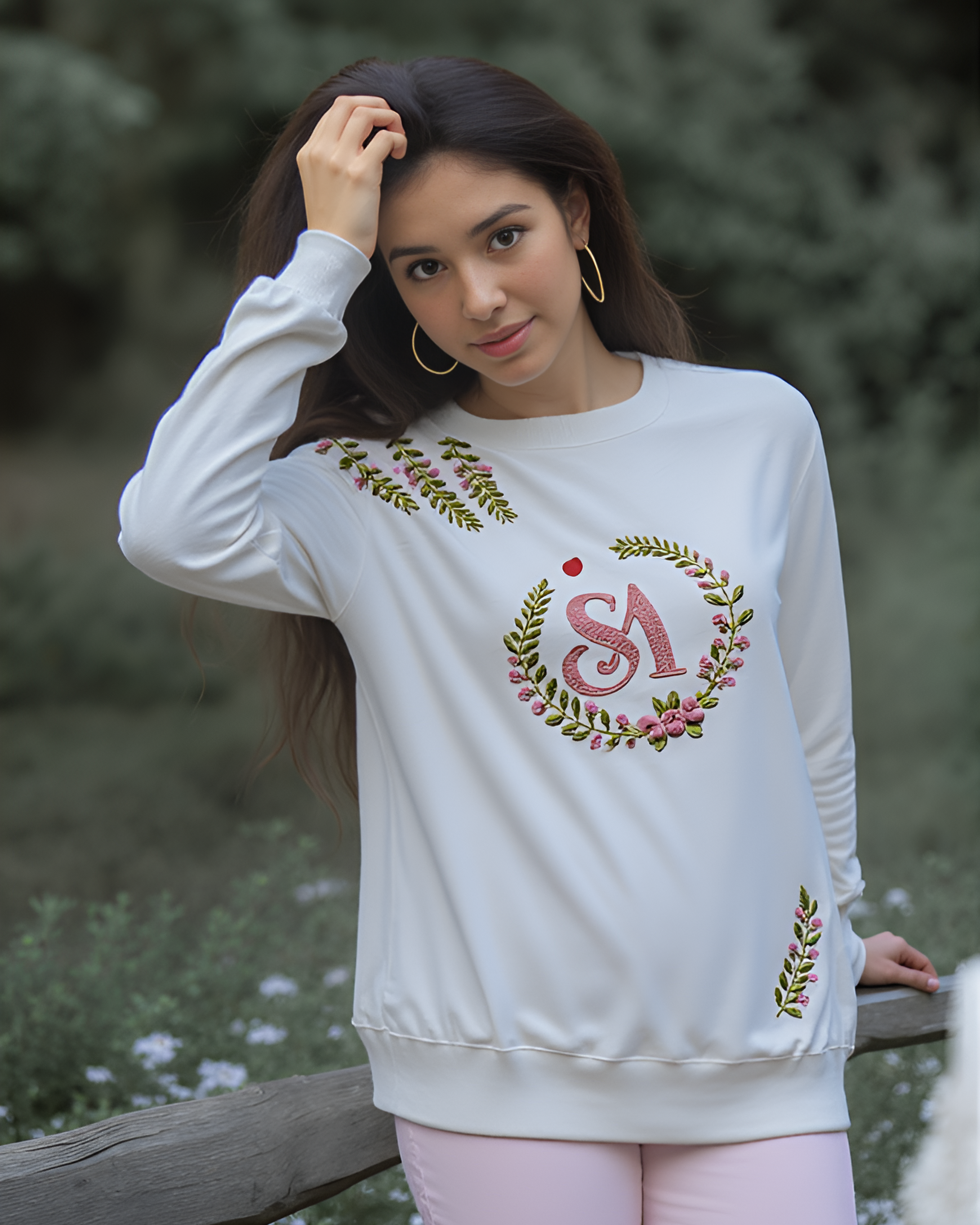 Woman wearing a white sweatshirt with a logo embroidered , standing outdoors with greenery in the background.