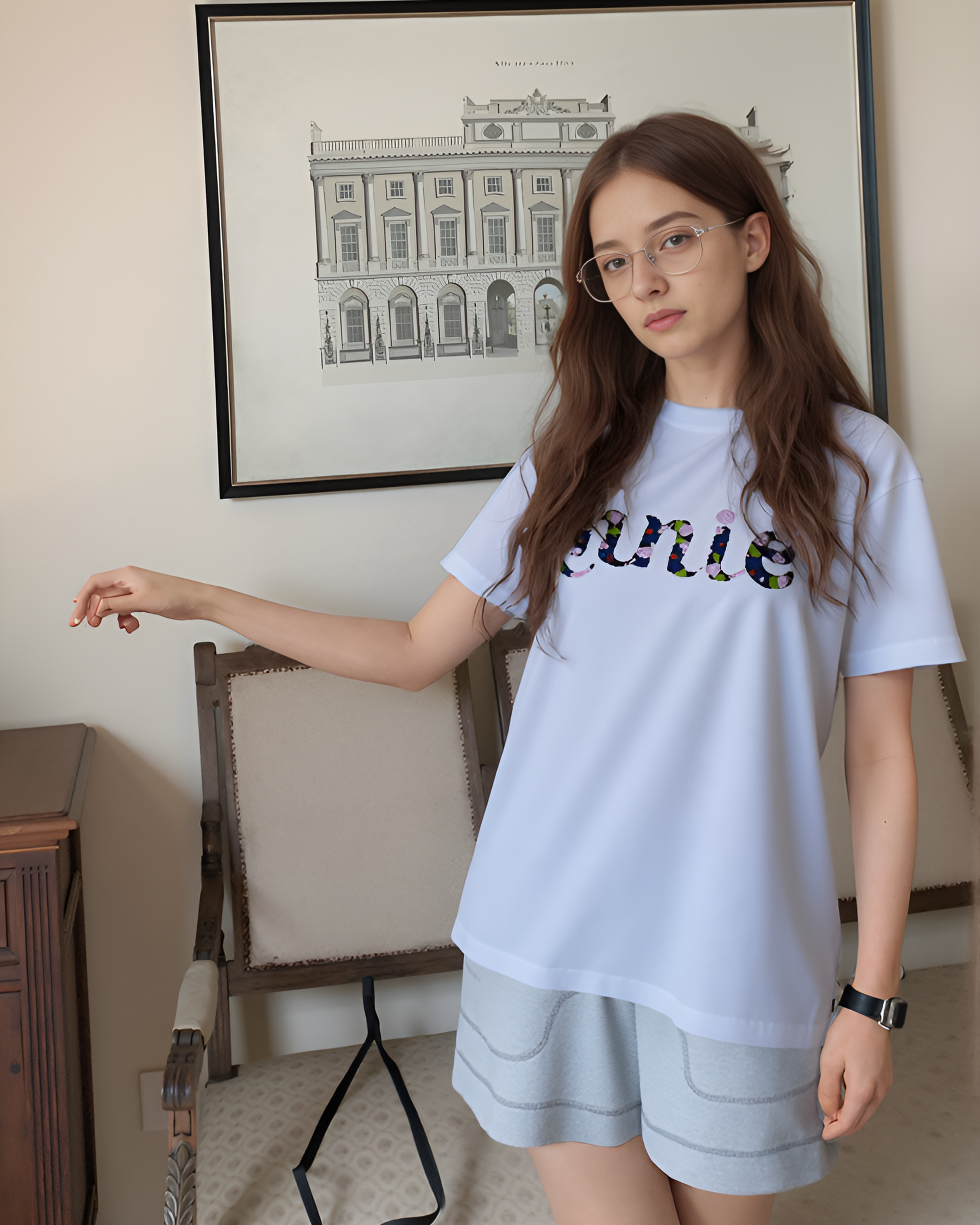 Woman wearing a white t-shirt with colorful embroidered custom name in a room with a chair and framed picture on the wall.