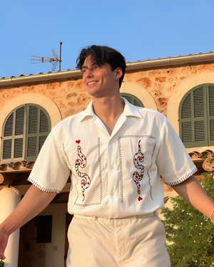 Man wearing a white embroidered shirt standing outdoors with a building and clear sky in the background