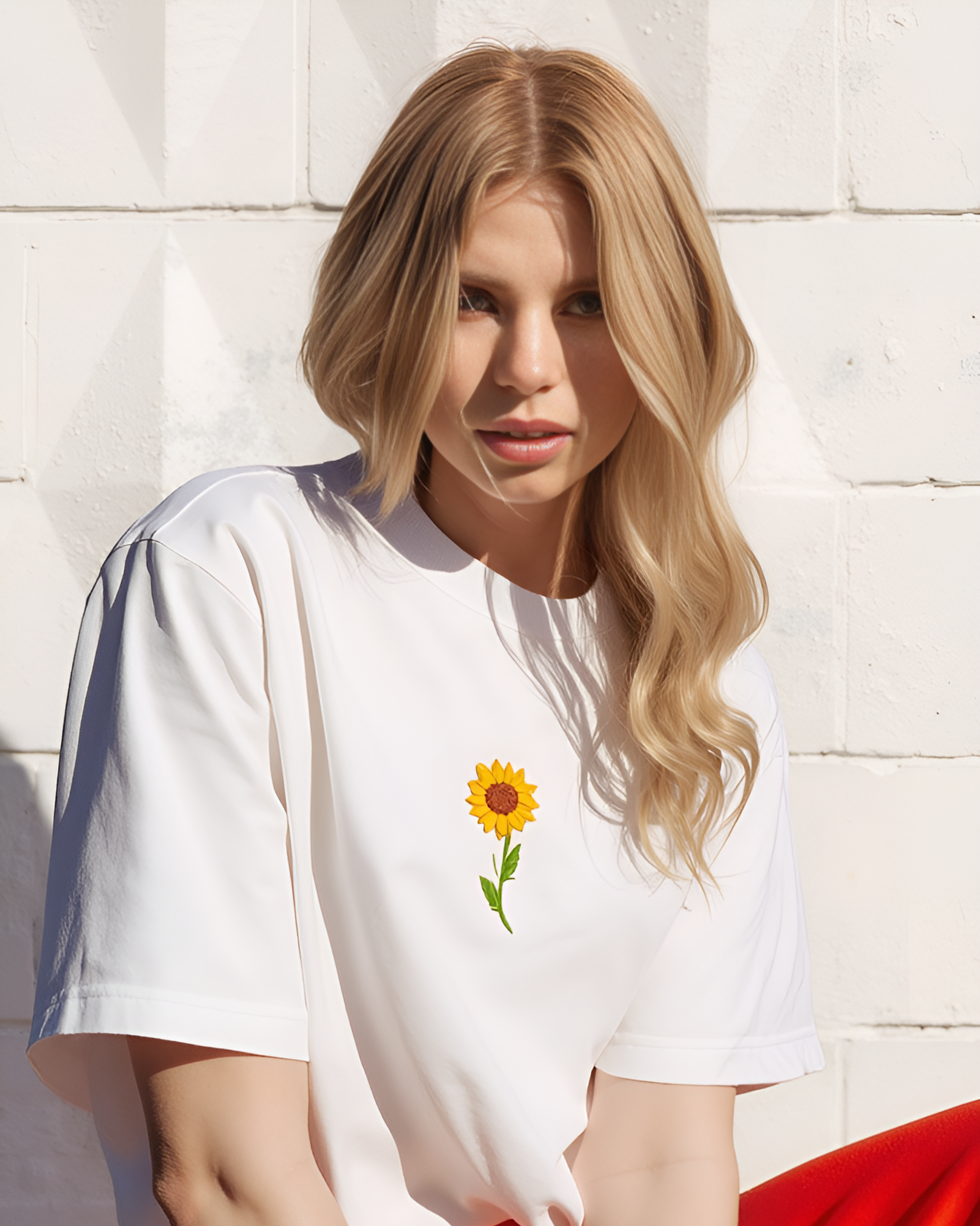 Woman wearing a white t-shirt with a sunflower design hand embroidered against a light-colored wall.