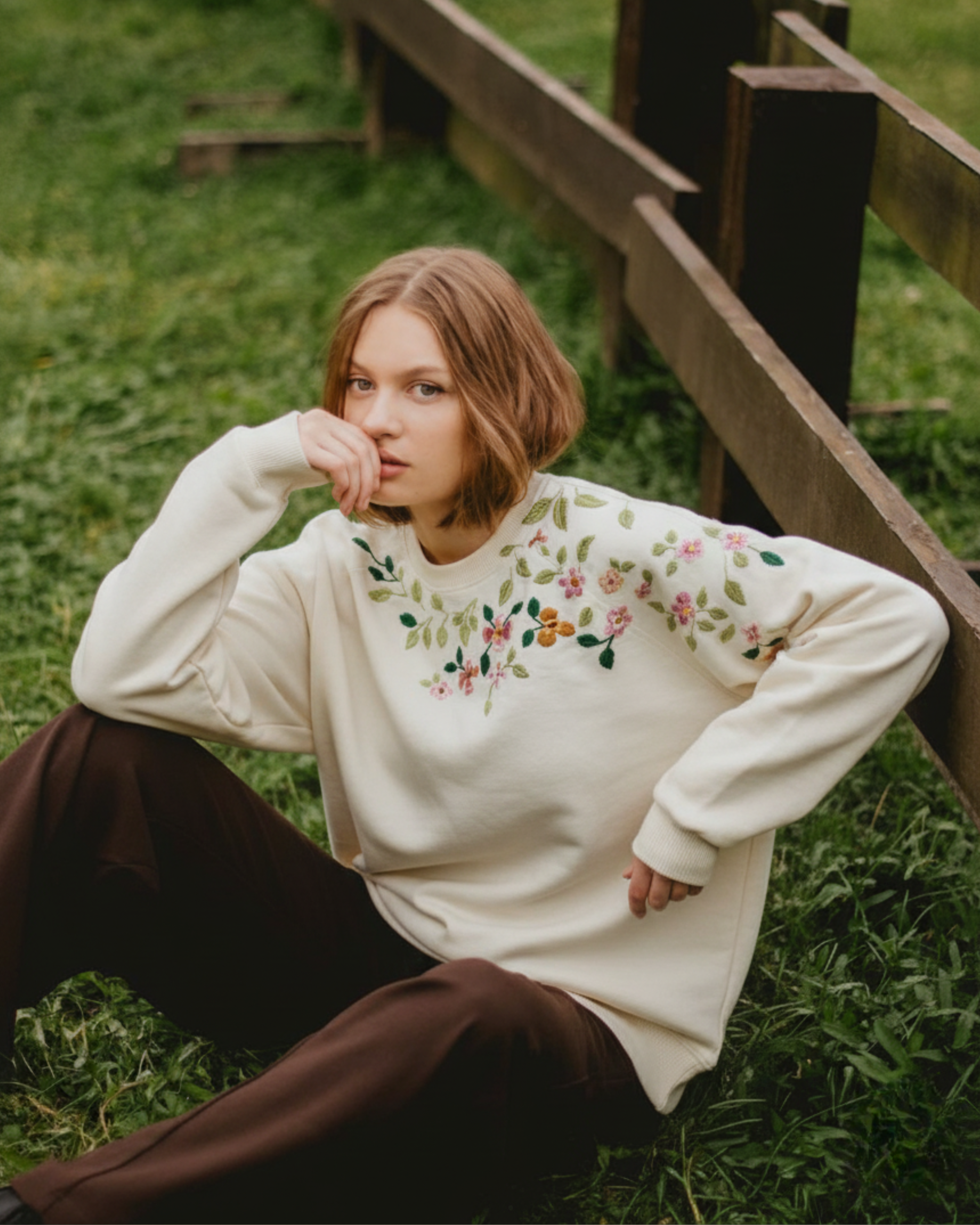 Woman wearing a cream sweater with floral embroidery sitting on grass next to a wooden fence.