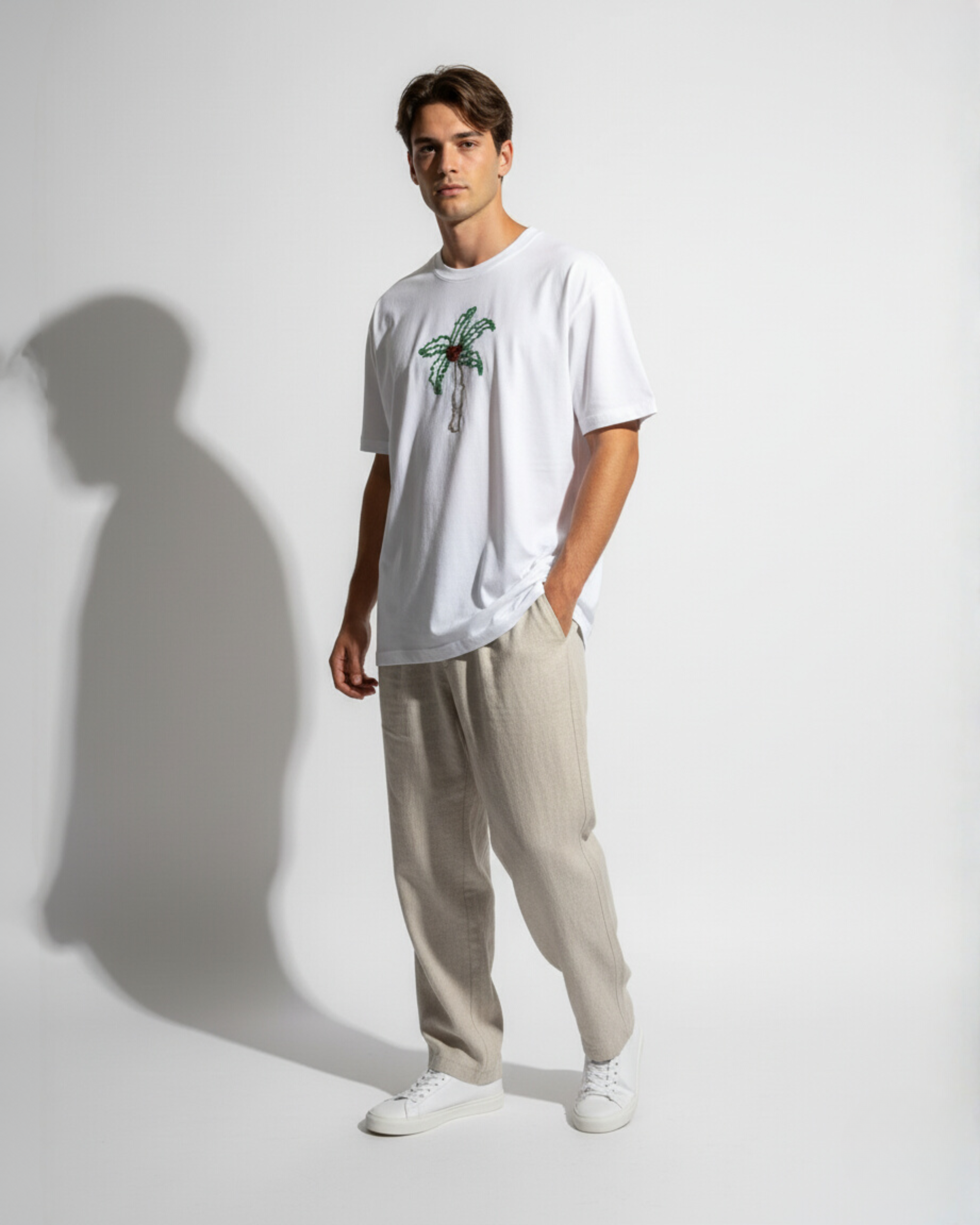 Man wearing a white t-shirt with a bead embroidered design and beige pants against a plain background