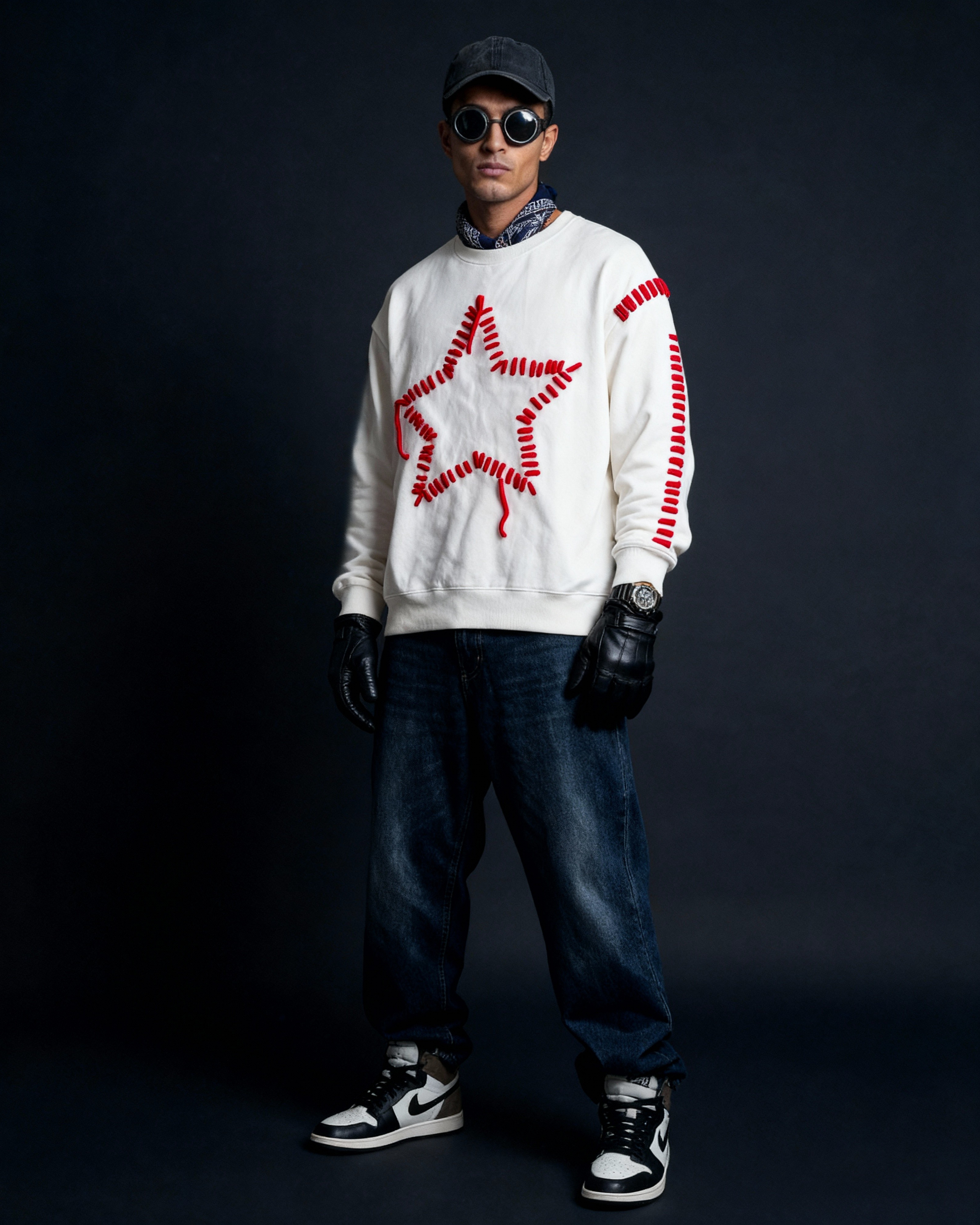 Person wearing a white sweater with red star design, dark jeans, black gloves, and black cap on a dark background