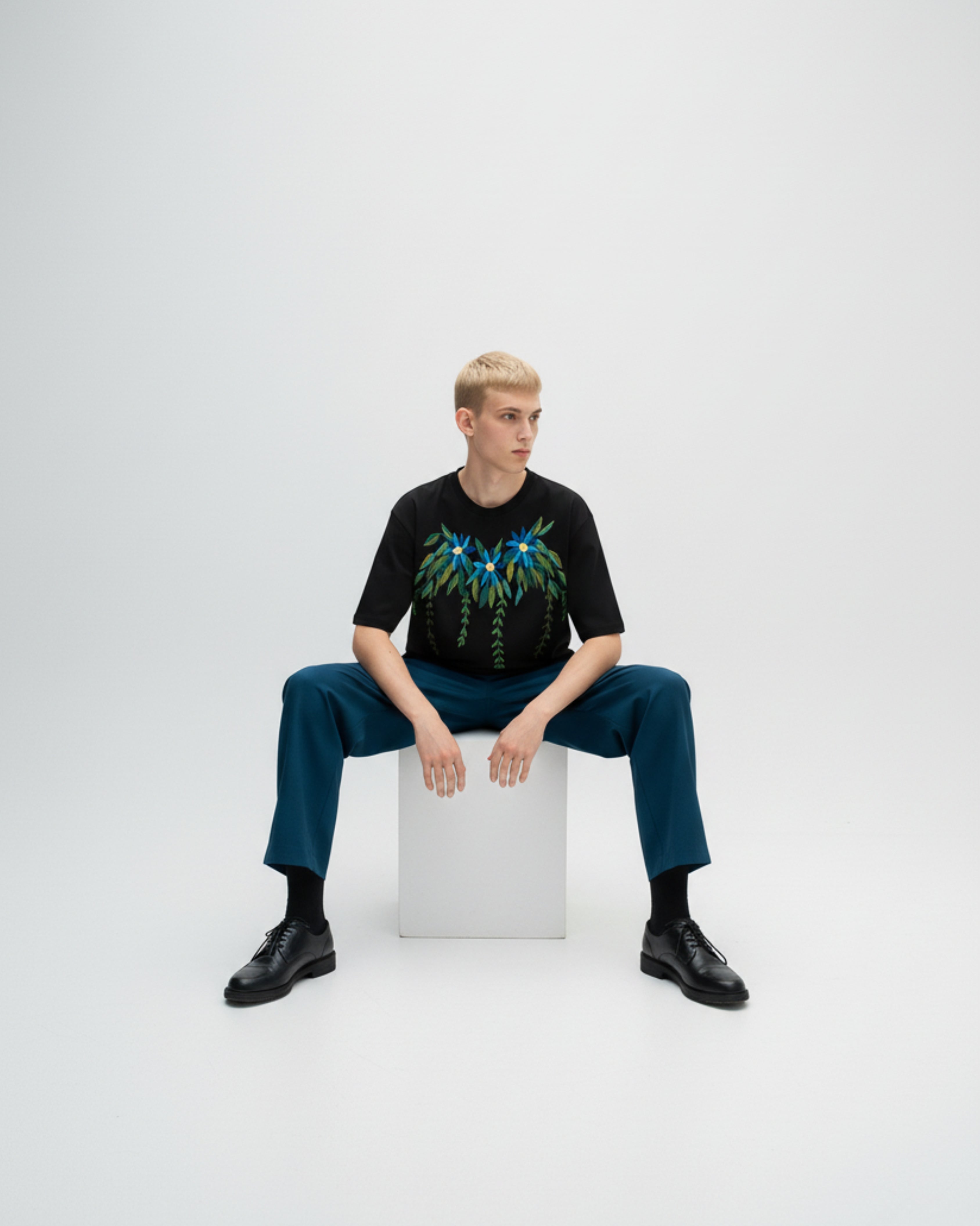 Person sitting on a white cube wearing a black t-shirt with a colorful hand embroidered design and blue pants.