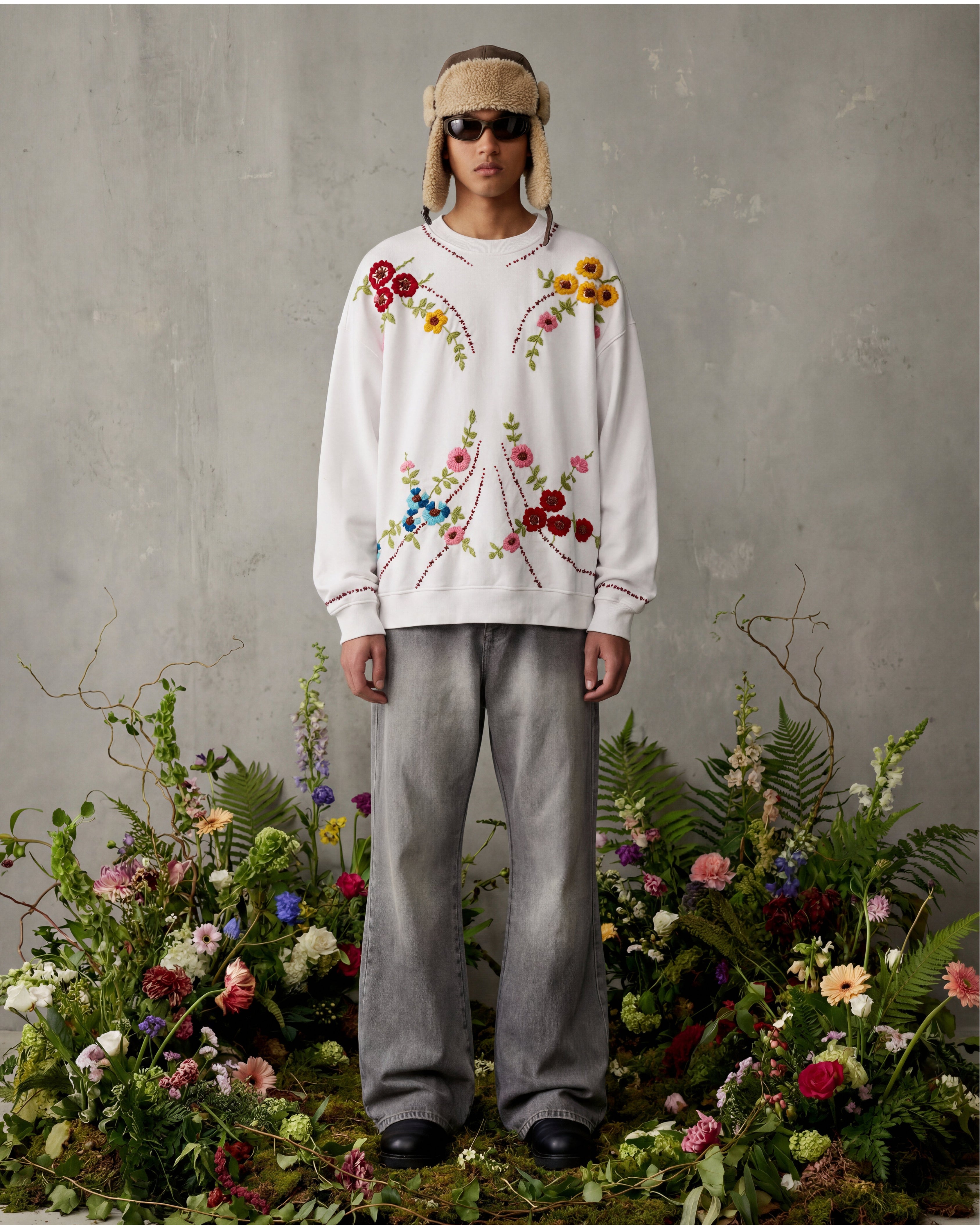 Person wearing a floral embroidered sweater and gray pants standing among flowers and plants.