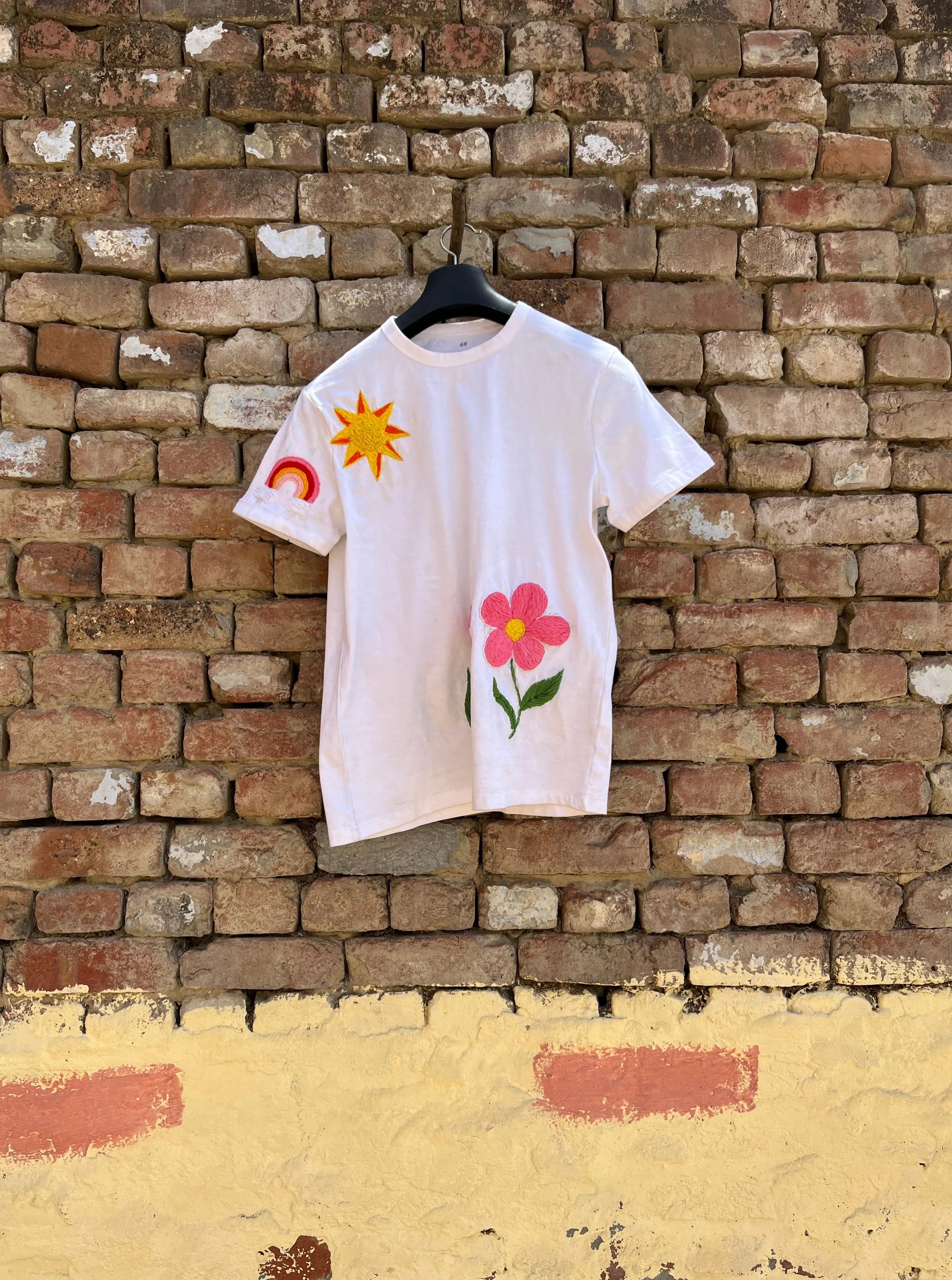 White t-shirt with colorful designs embroidered on a hanger against a brick wall.
