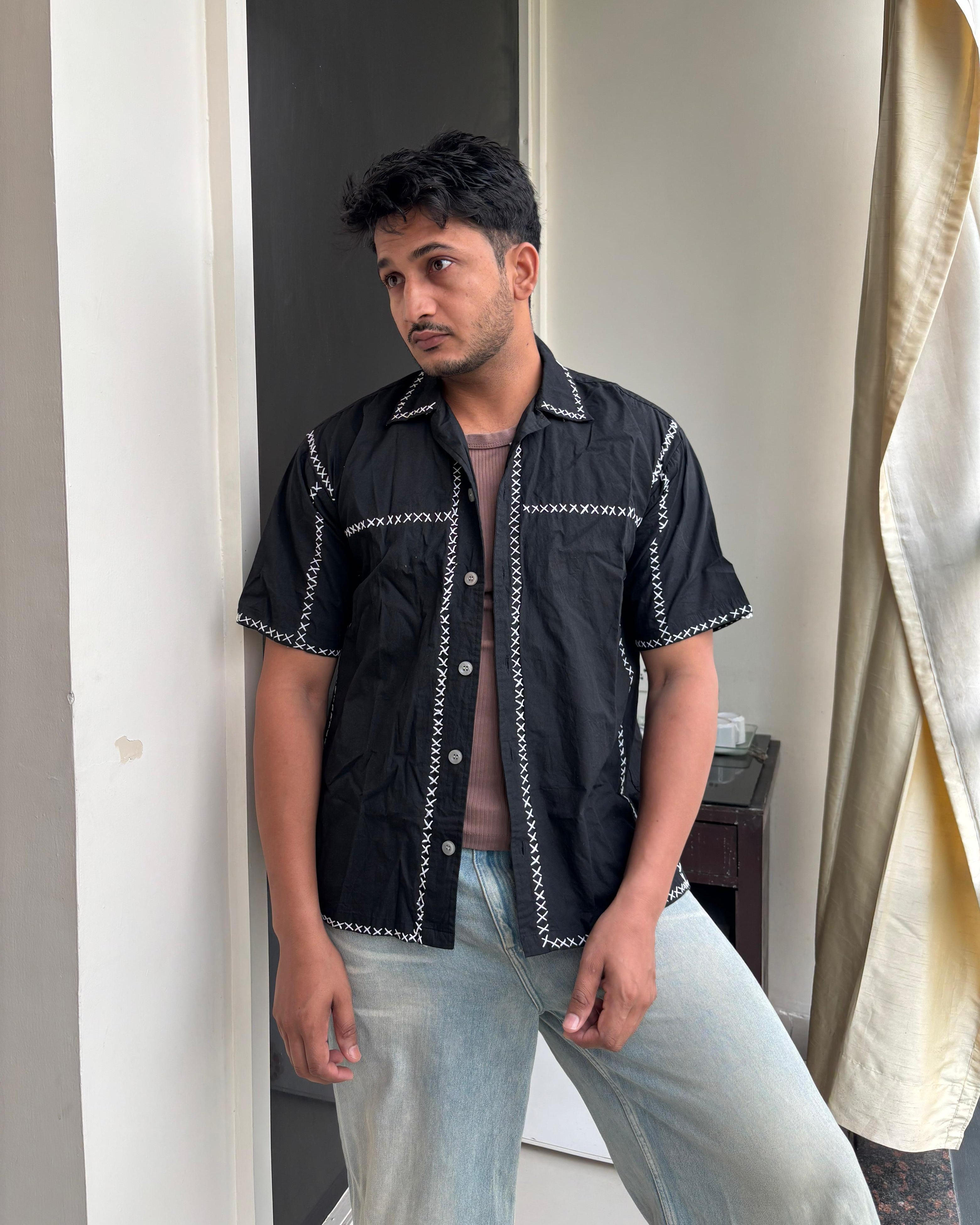A man wearing a black short-sleeved shirt with hand-embroidered X-pattern and a vintage-inspired Cuban collar.