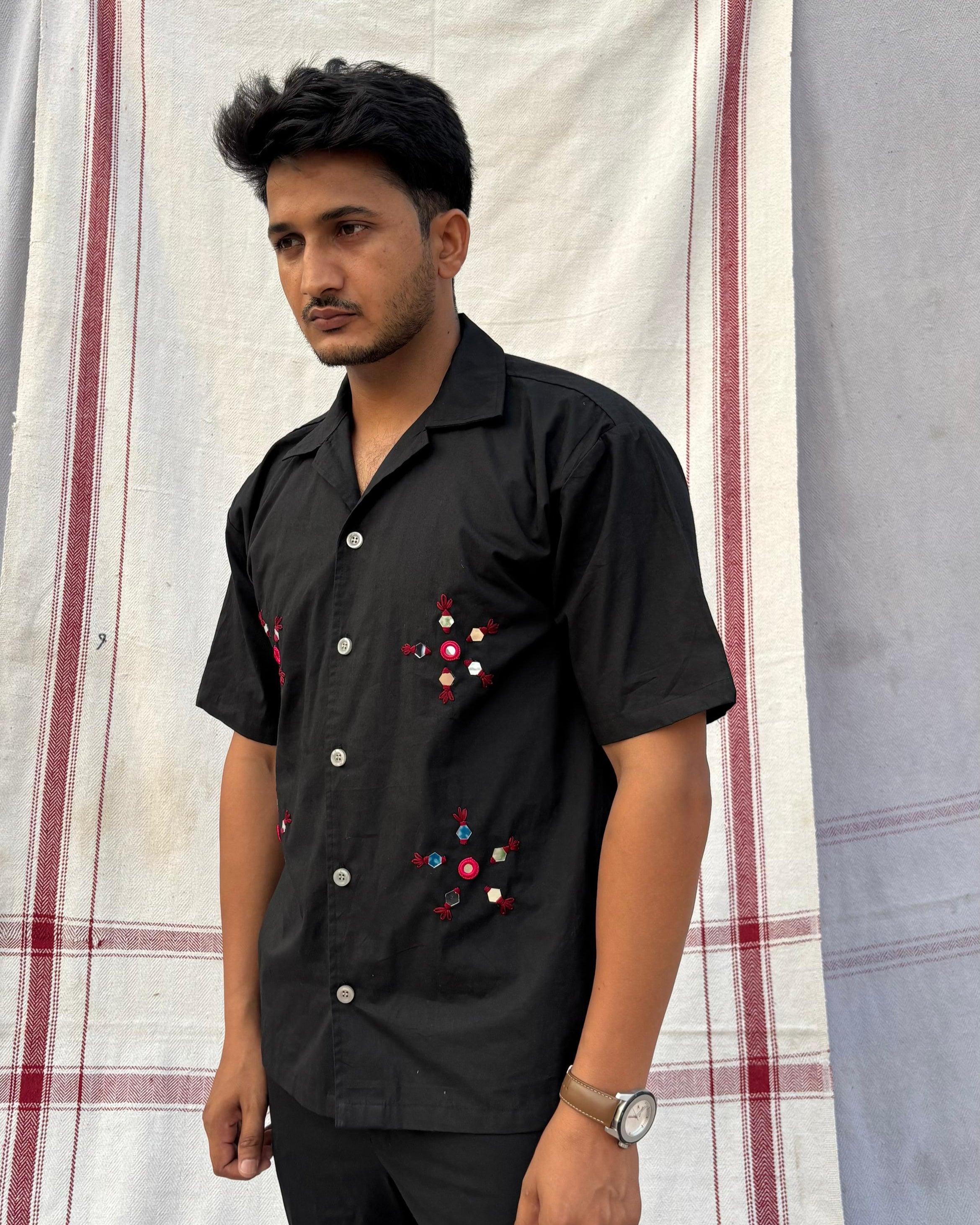 Person wearing a mirror work embroidered black shirt in front of a striped fabric background