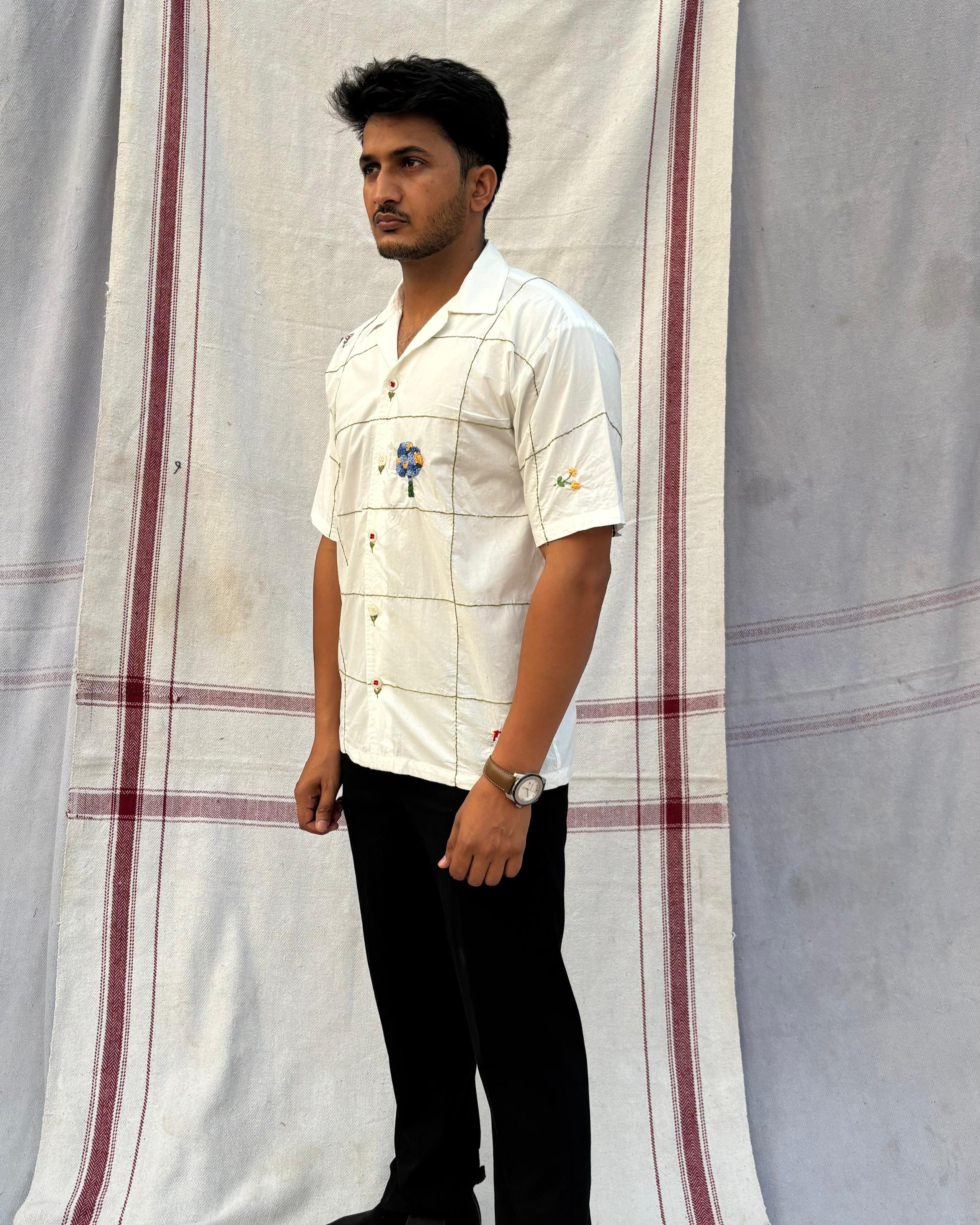 A man standing against a striped backdrop, wearing a white short-sleeved cotton shirt with embroidered floral and insect patterns and a Cuban collar.