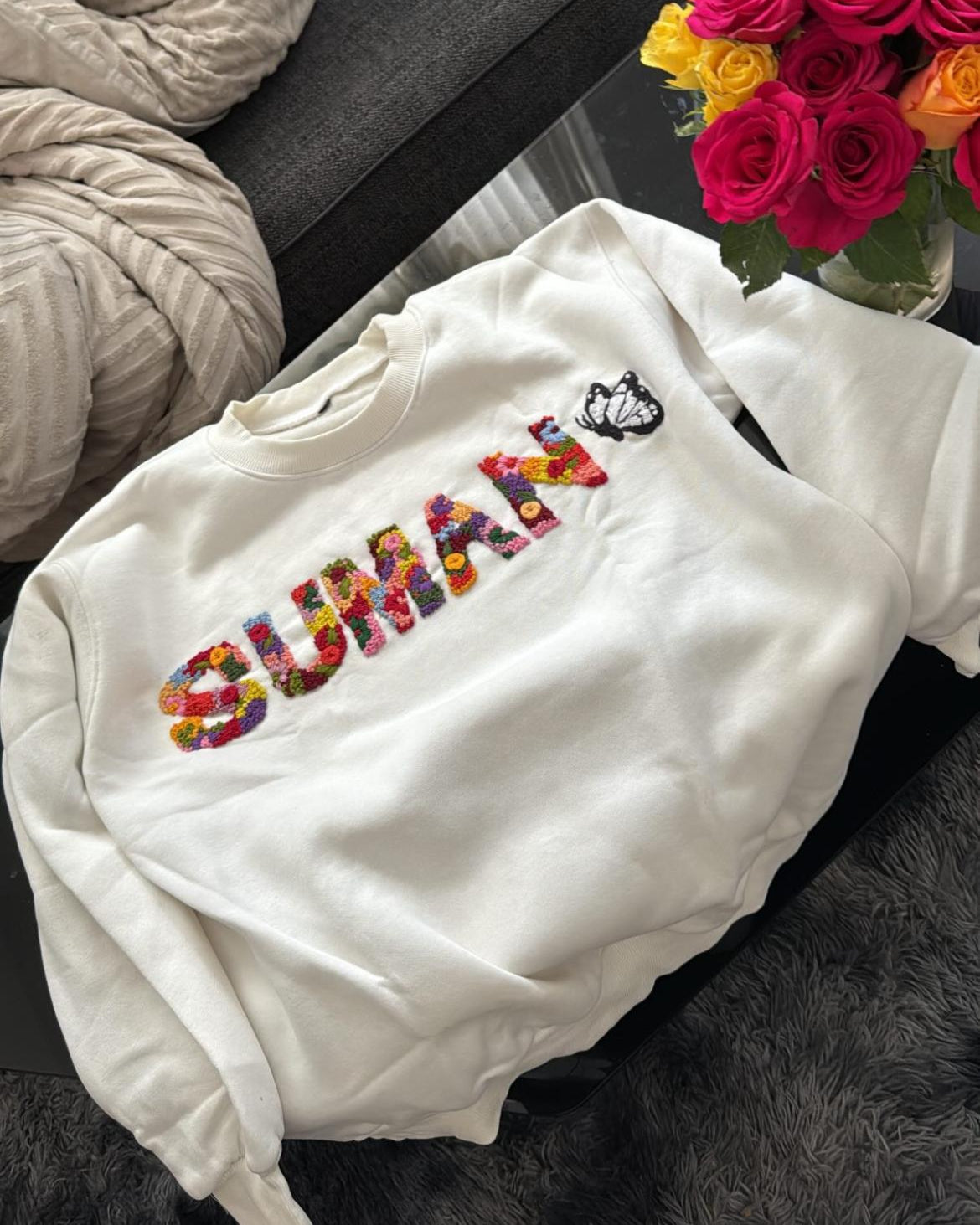 Flatlay of custom name sweatshirt in beige with soft embroidery, surrounded by candles and gift ribbon for a cozy gifting vibe on a dark surface with flowers in the background