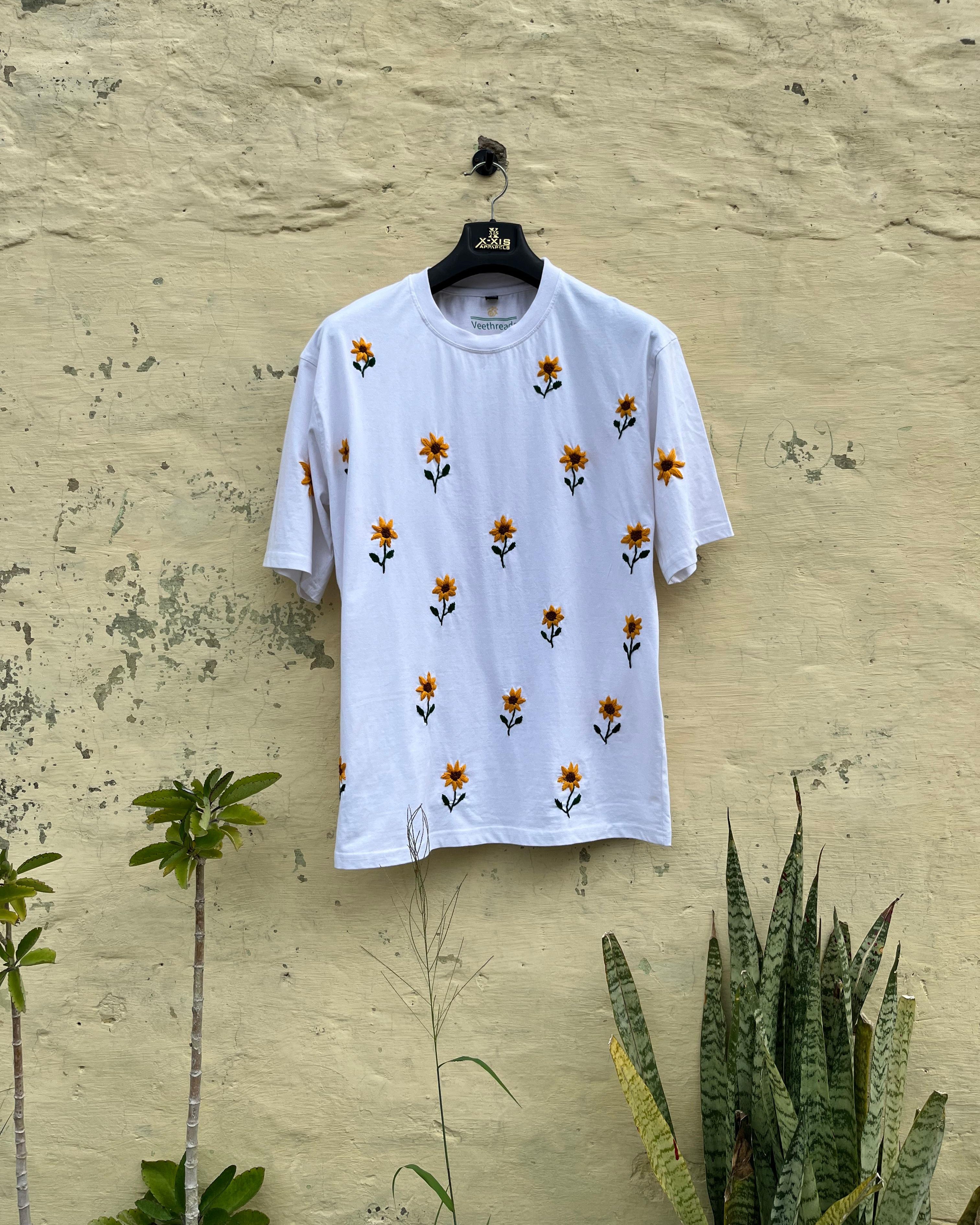 White t-shirt with floral patterns on a hanger against a textured beige wall.