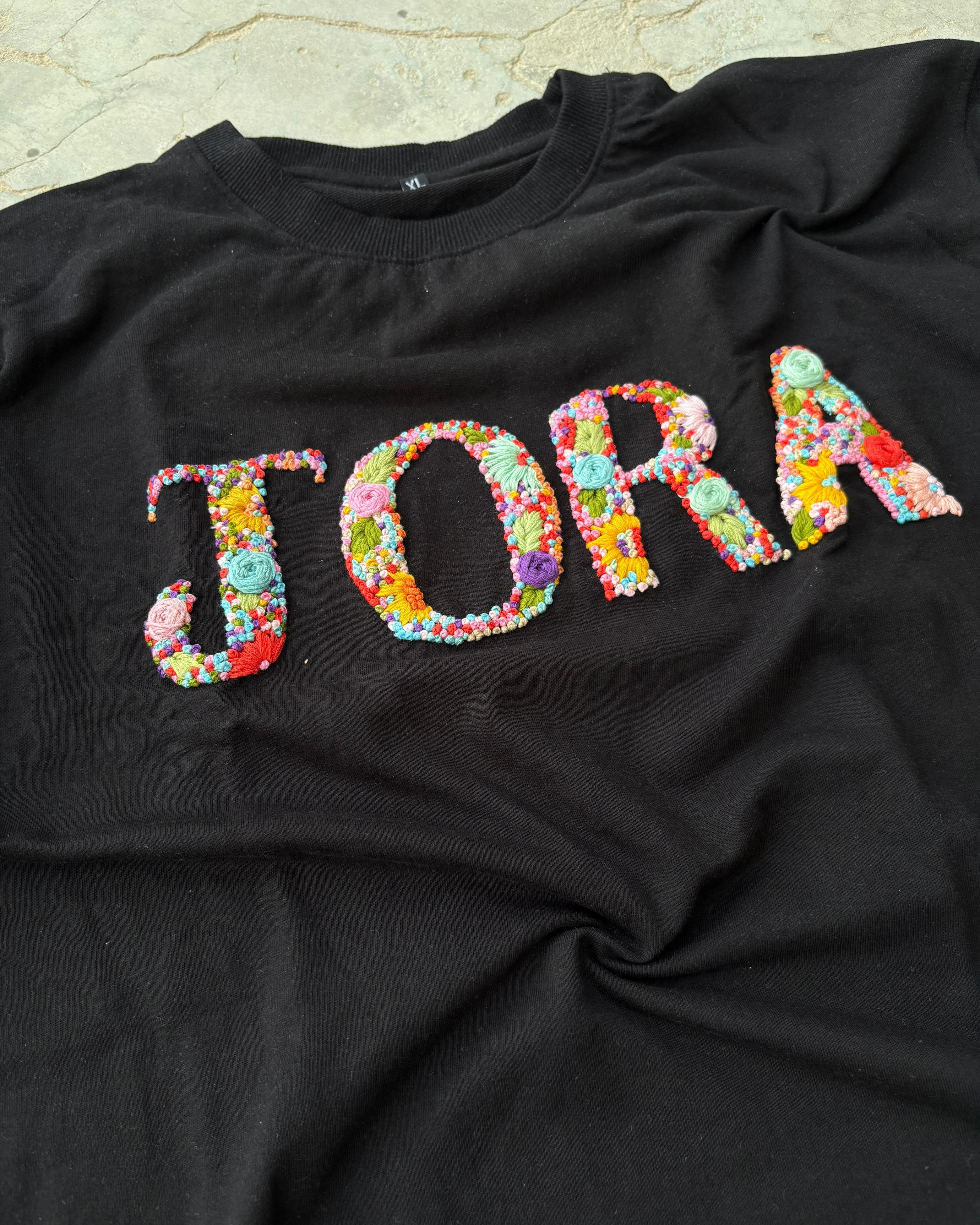 Custom t-shirt printing with name detail in multicolored thread, embroidered by skilled artisans on high-GSM cotton.